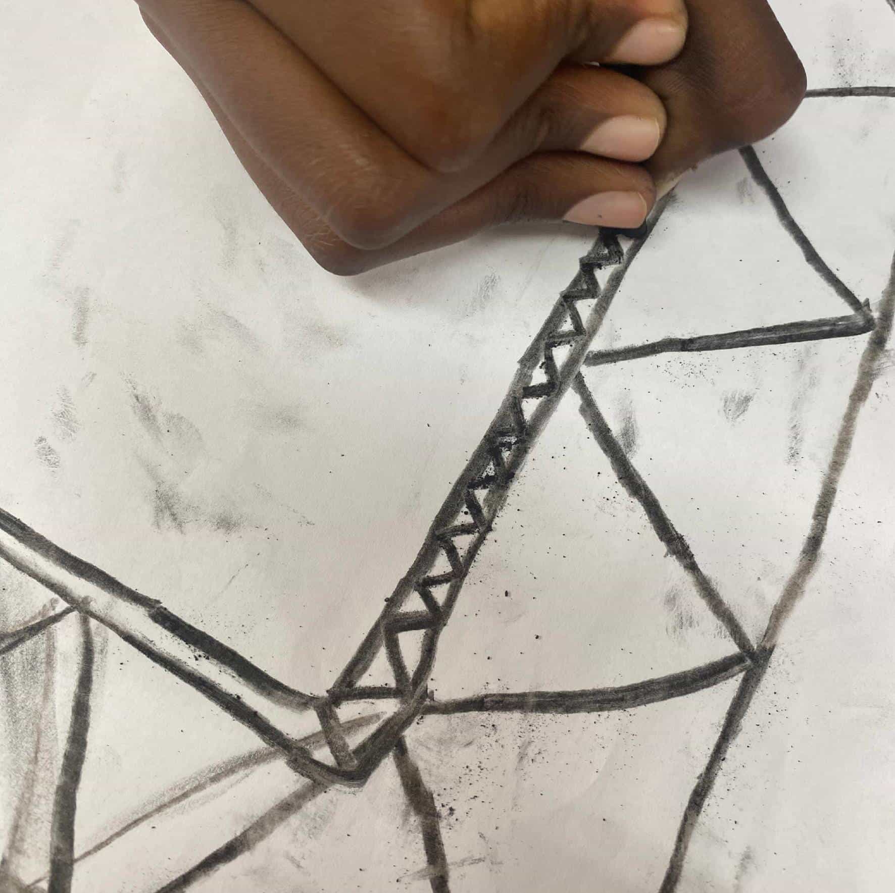A hand drawing geometric shapes with black charcoal on white paper, reminiscent of a Gogo Mahlangu Legacy Project. The hand adds triangular patterns inside a thick outlined shape; charcoal smudges mark the Celebration of creative expression.