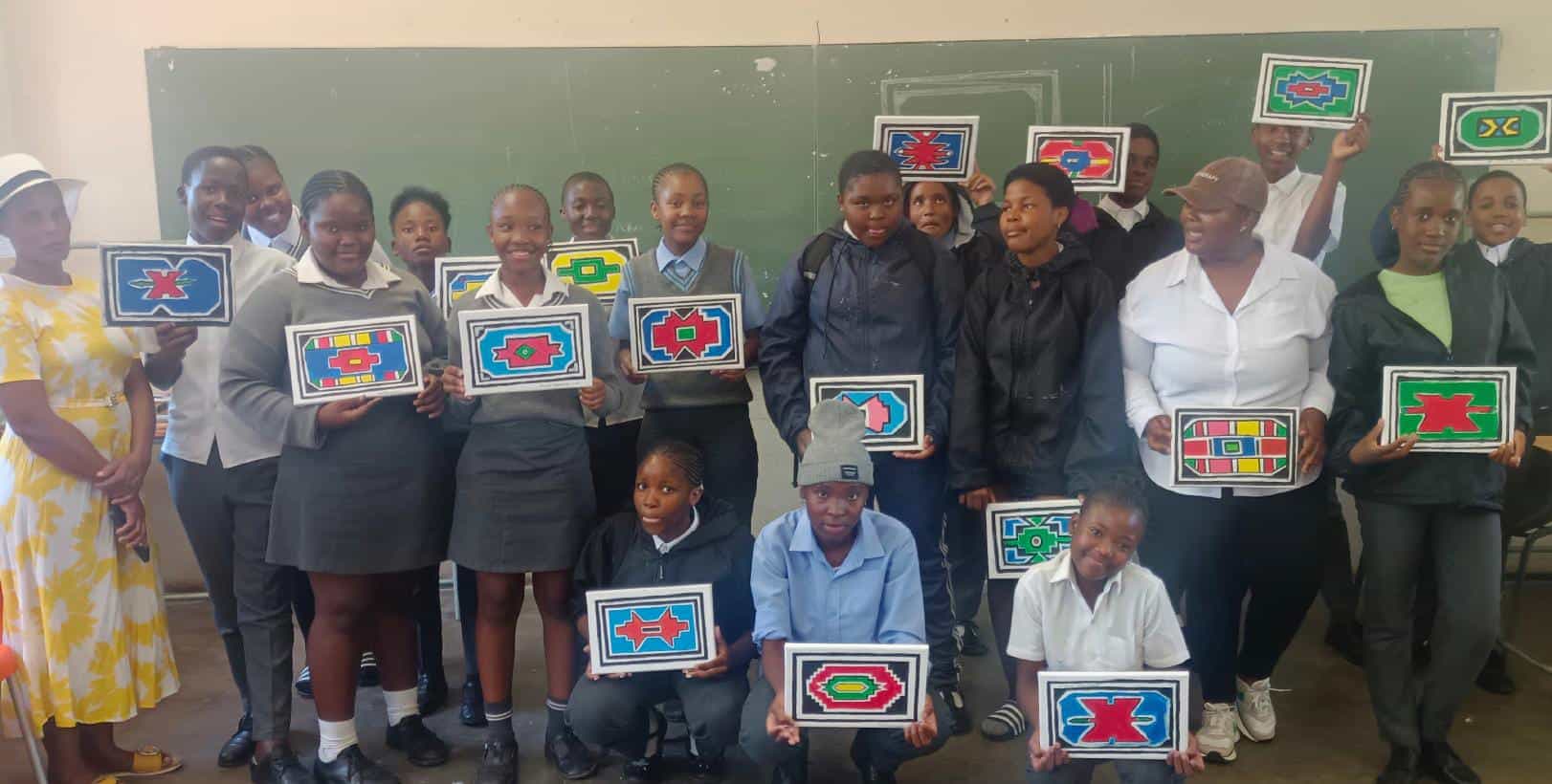 A group of students and two adults stand in a classroom, smiling and holding colourful, rectangular artworks with geometric patterns—a vibrant Celebration for the Legacy Project—posing together in front of a green blackboard.