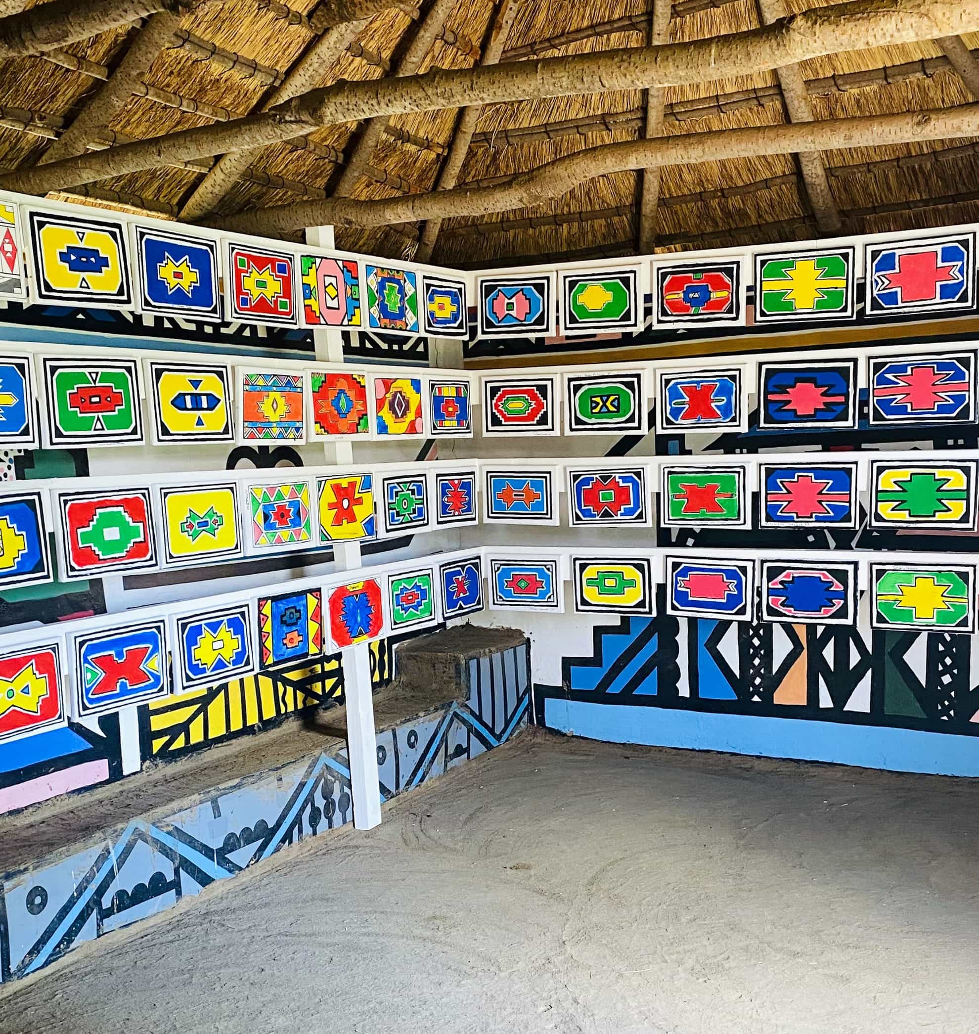 A thatched-roof hut with walls decorated in rows of colourful, geometric Ndebele patterns—a Celebration of Gogo Mahlangu’s artistry. The sandy floor contrasts with the vivid wall art, honouring the Legacy Project’s vibrant heritage.