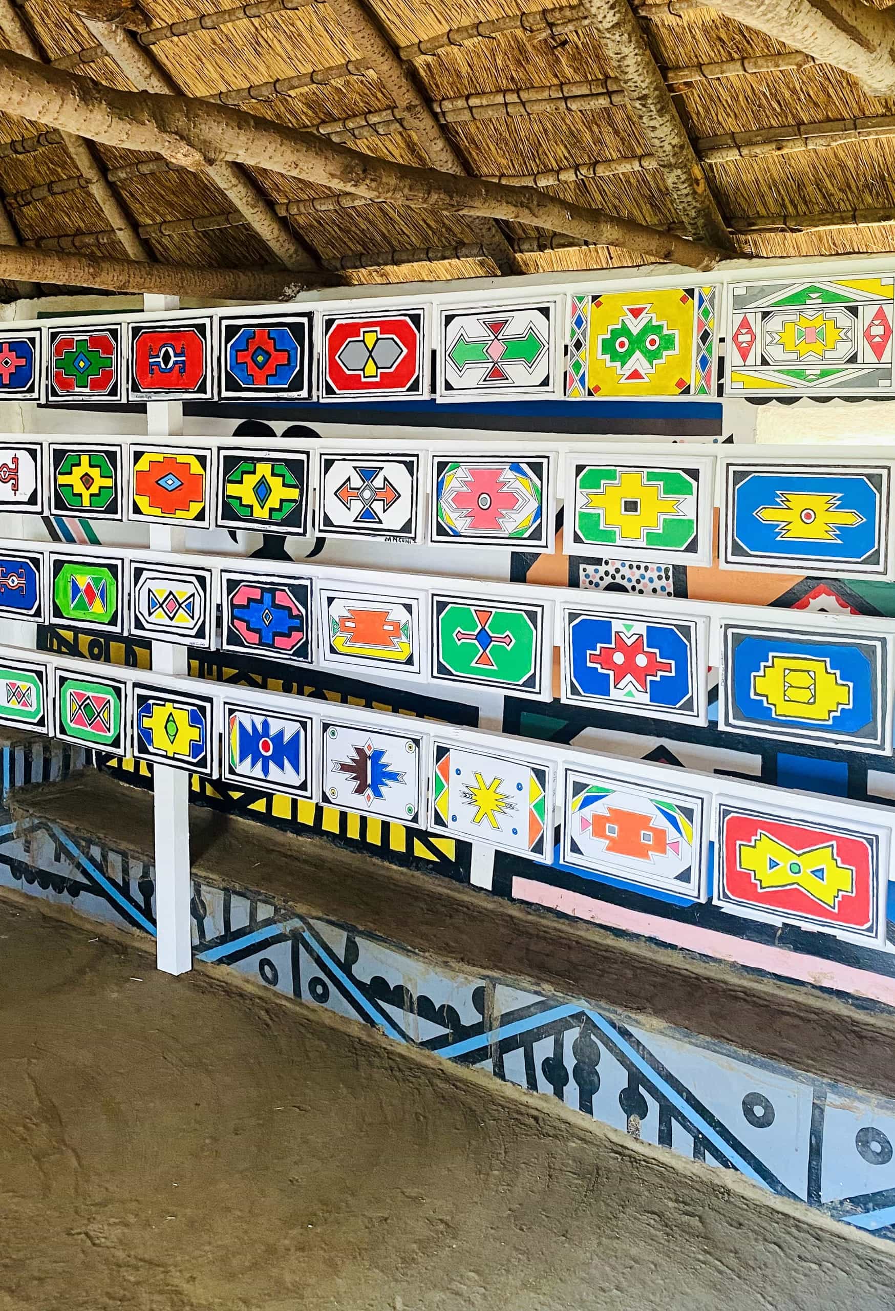 A wall lined with colourful rectangular panels painted in vibrant geometric Ndebele patterns, part of the Legacy Project celebrating Gogo Mahlangu. The floor is brown earth, and the lower wall features striking geometric designs in black, white, and blue.