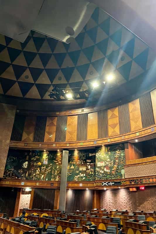 A legislative chamber with wooden desks and chairs arranged in rows. The walls feature colourful mural art and wooden panels with geometric designs. The ceiling is decorated with a triangular pattern. Overhead lights illuminate the space.