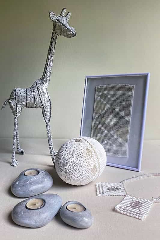 A beaded giraffe figurine, a framed geometric beadwork, a white beaded egg, three smooth stone tealight holders, and two beaded necklaces are arranged on a neutral surface against a light background.