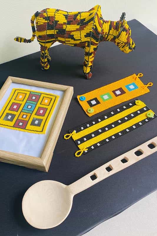 A beaded cow sculpture, a framed geometric beadwork, three colourful beaded bracelets, and a large wooden spoon with square holes are displayed on a black surface against a light background.