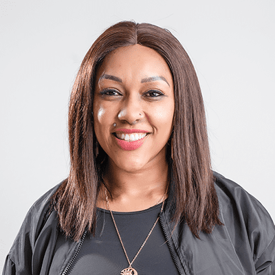 A woman with straight, shoulder-length brown hair smiles at the camera. She wears a black top, a black jacket, and a gold necklace. Her makeup is natural, and she has a nose piercing. The background is plain and light-colored.