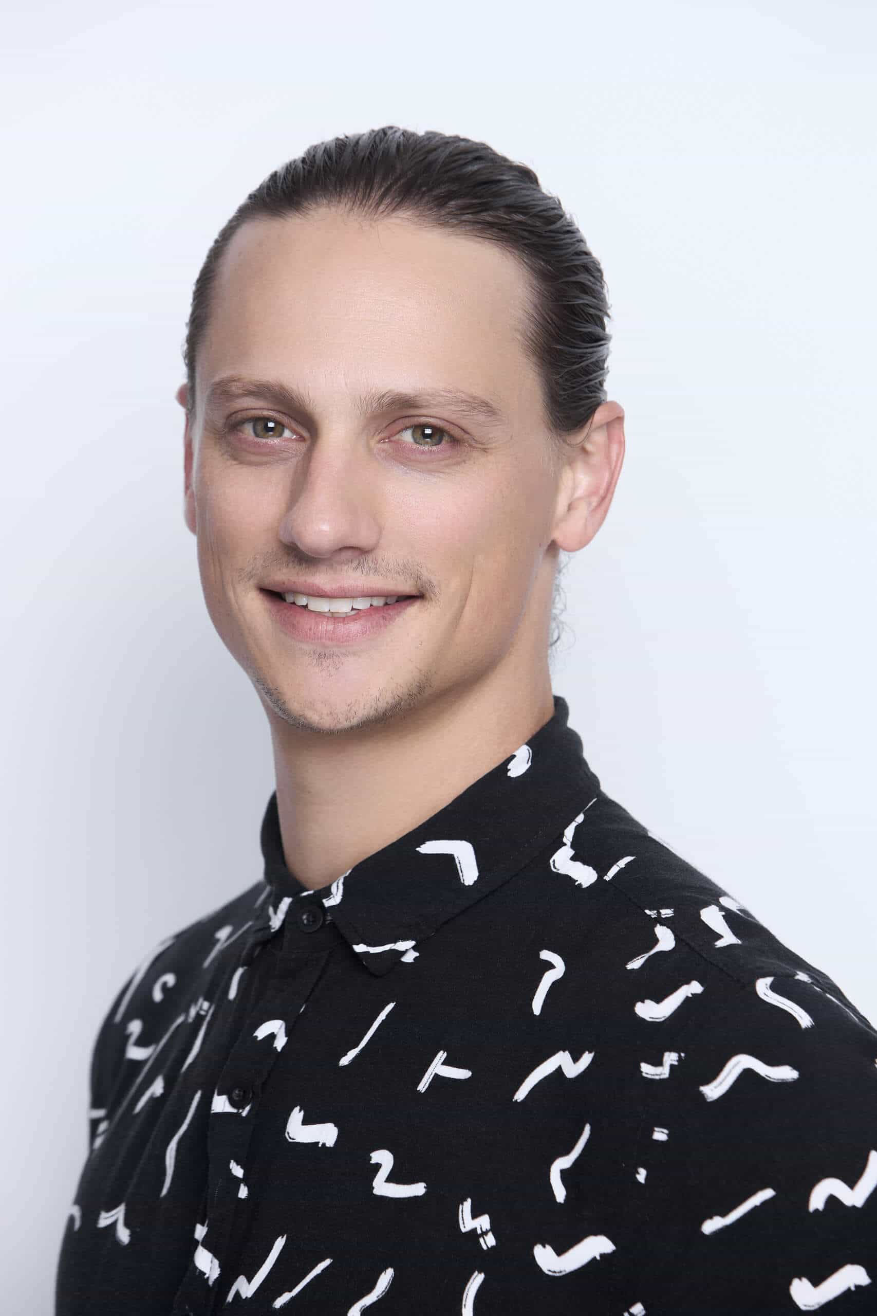 A person with light skin and light brown hair pulled back is smiling slightly at the camera. They are wearing a black button-up shirt with a white abstract squiggle pattern, and the background is plain white.