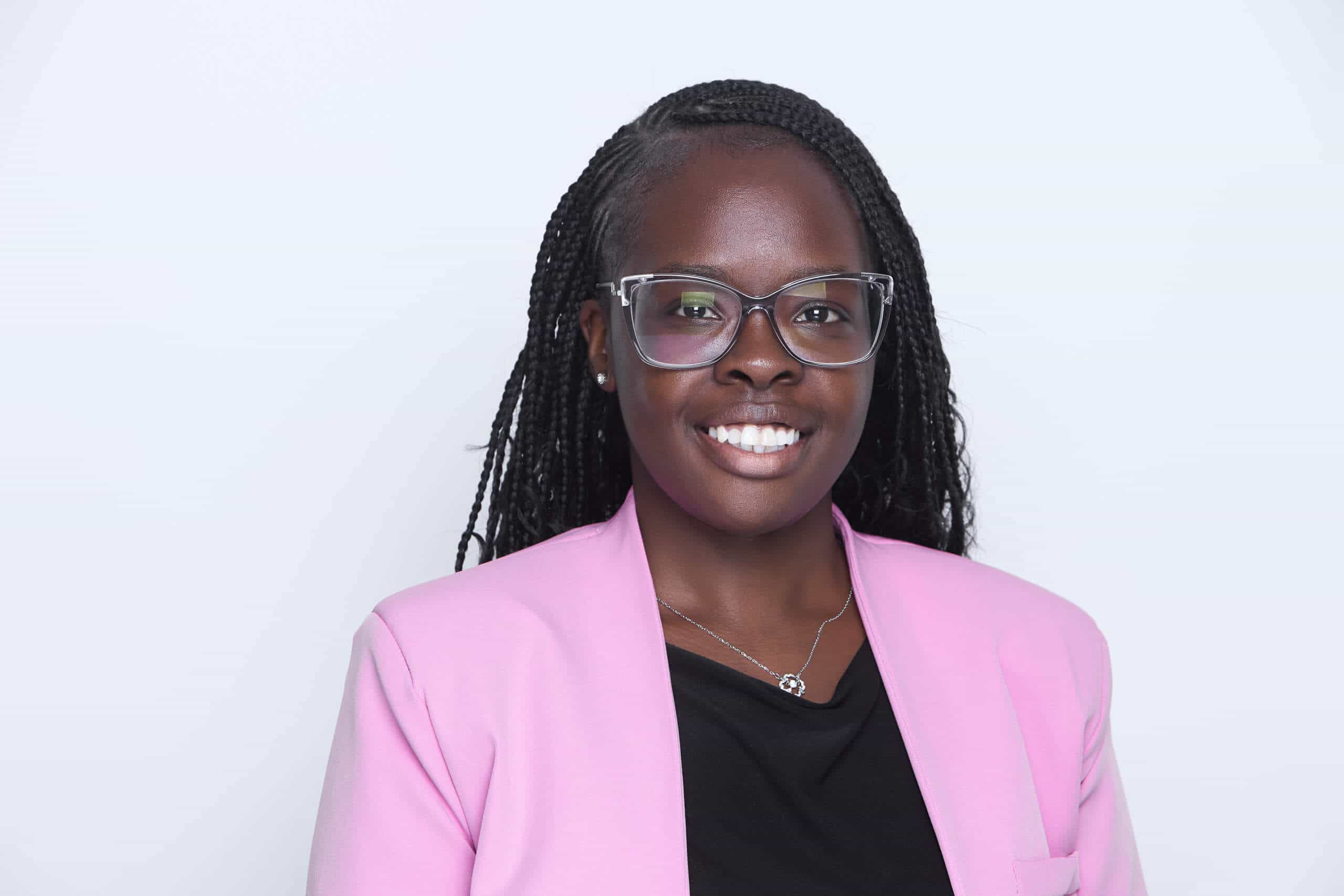 A woman with long braided hair, wearing glasses, a pink blazer over a black top, and a silver necklace, smiles at the camera against a plain white background.