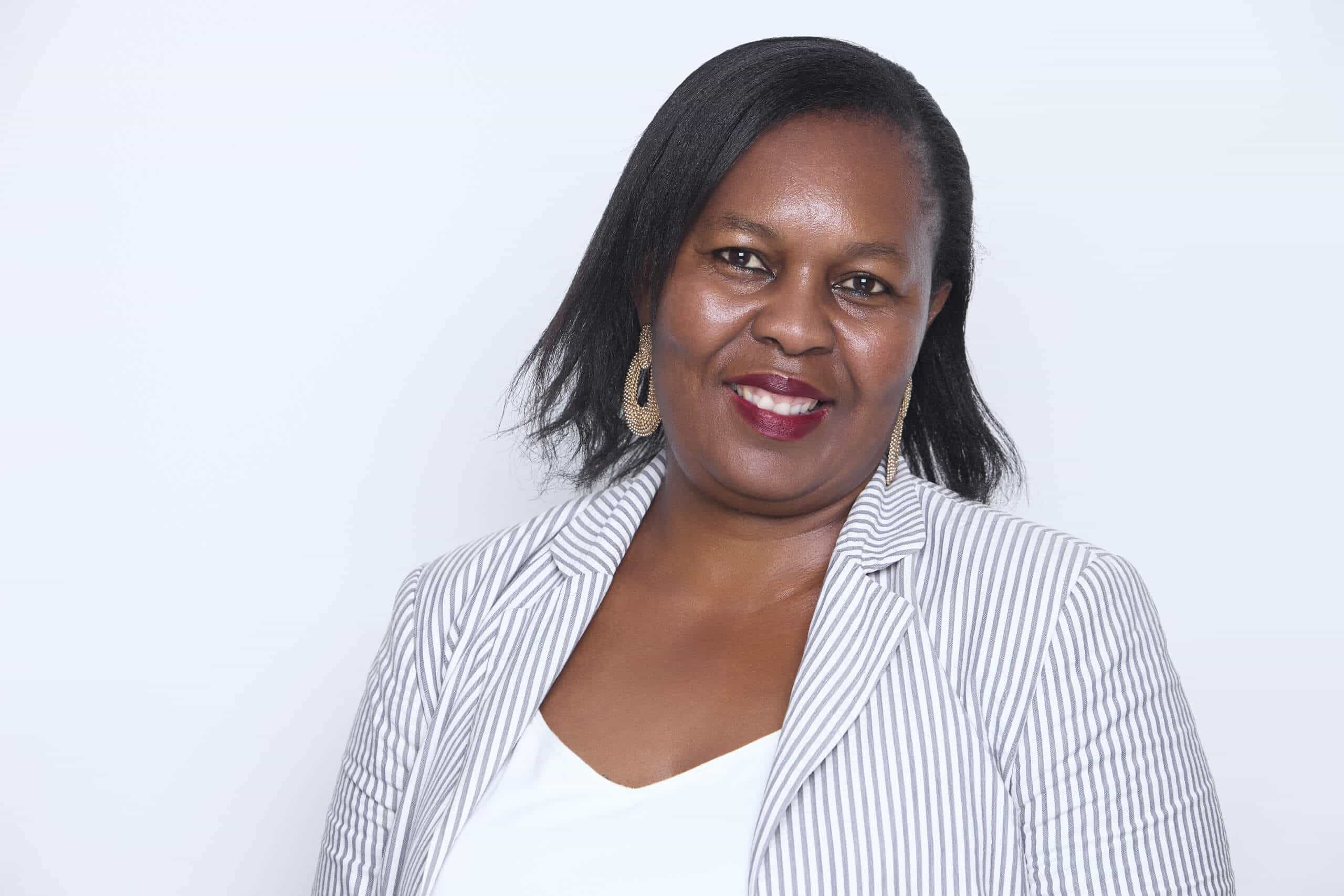 A woman with dark skin and straight black hair wears gold hoop earrings, a white top, and a light gray striped blazer. She is smiling and posed against a plain white background.