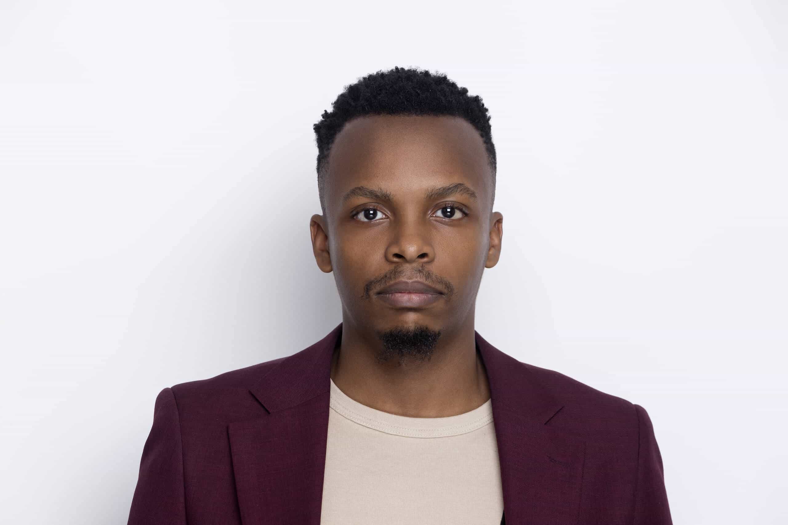 A man with short black hair and a goatee looks directly at the camera. He wears a burgundy blazer over a beige shirt and stands against a plain white background. His expression is neutral and calm.