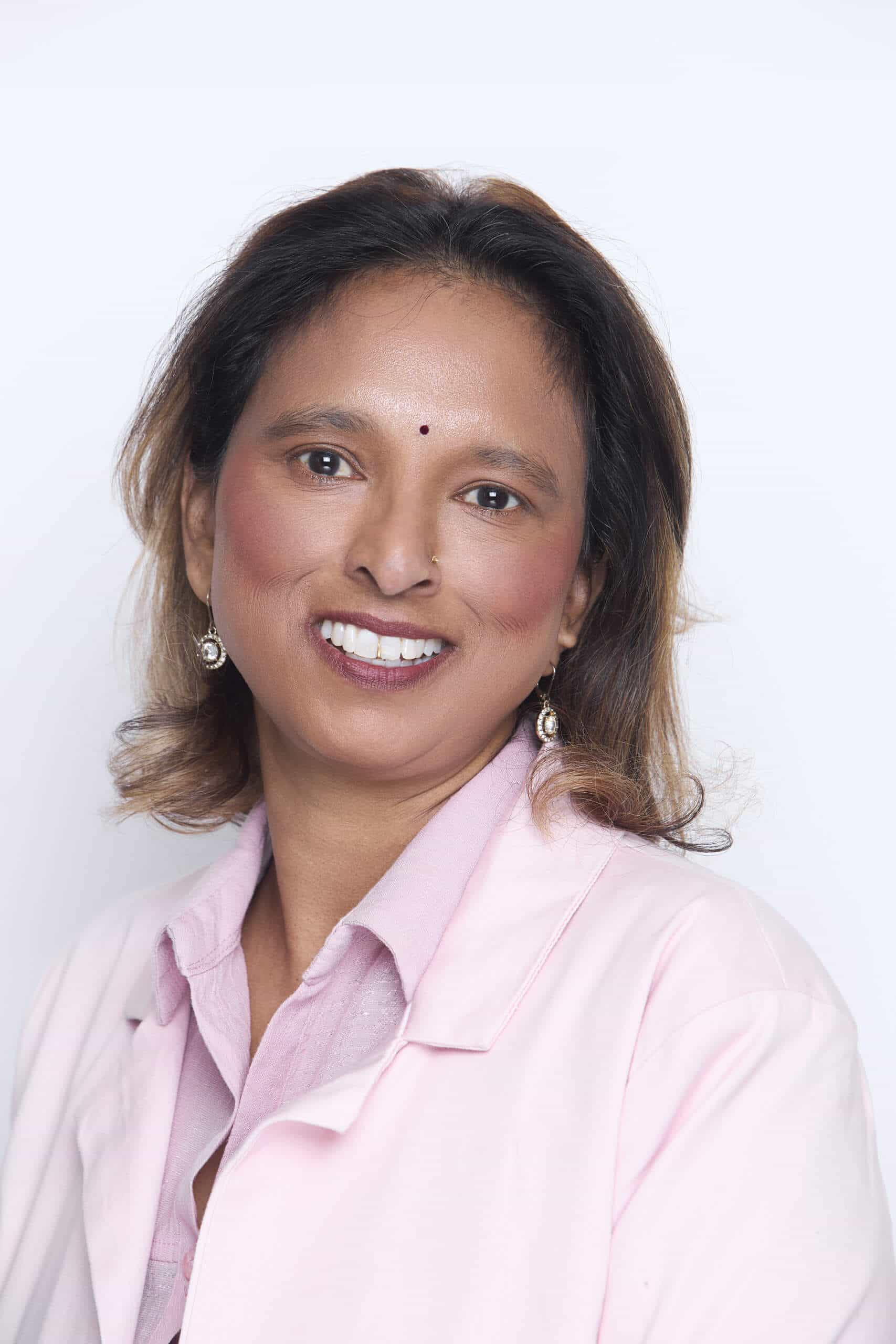 A woman wearing a light pink lab coat and matching collared shirt smiles at the camera. She has medium-length, wavy dark hair with light brown highlights, medium brown skin, and wears drop earrings. There is a small bindi on her forehead. The background is white.