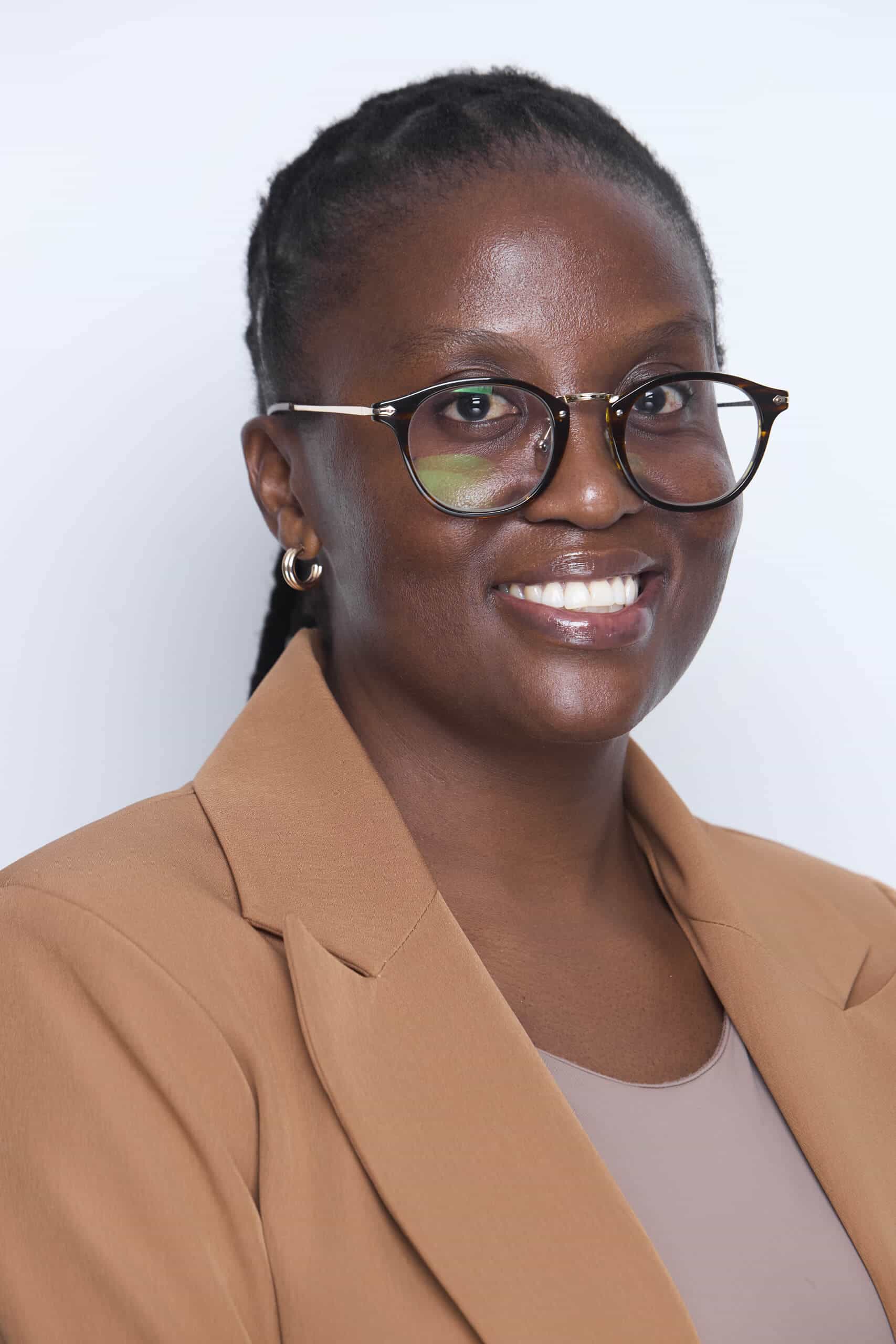 A woman with dark skin and braided hair, wearing round glasses, hoop earrings, a tan blazer, and a light mauve top, smiles at the camera against a plain, light background.