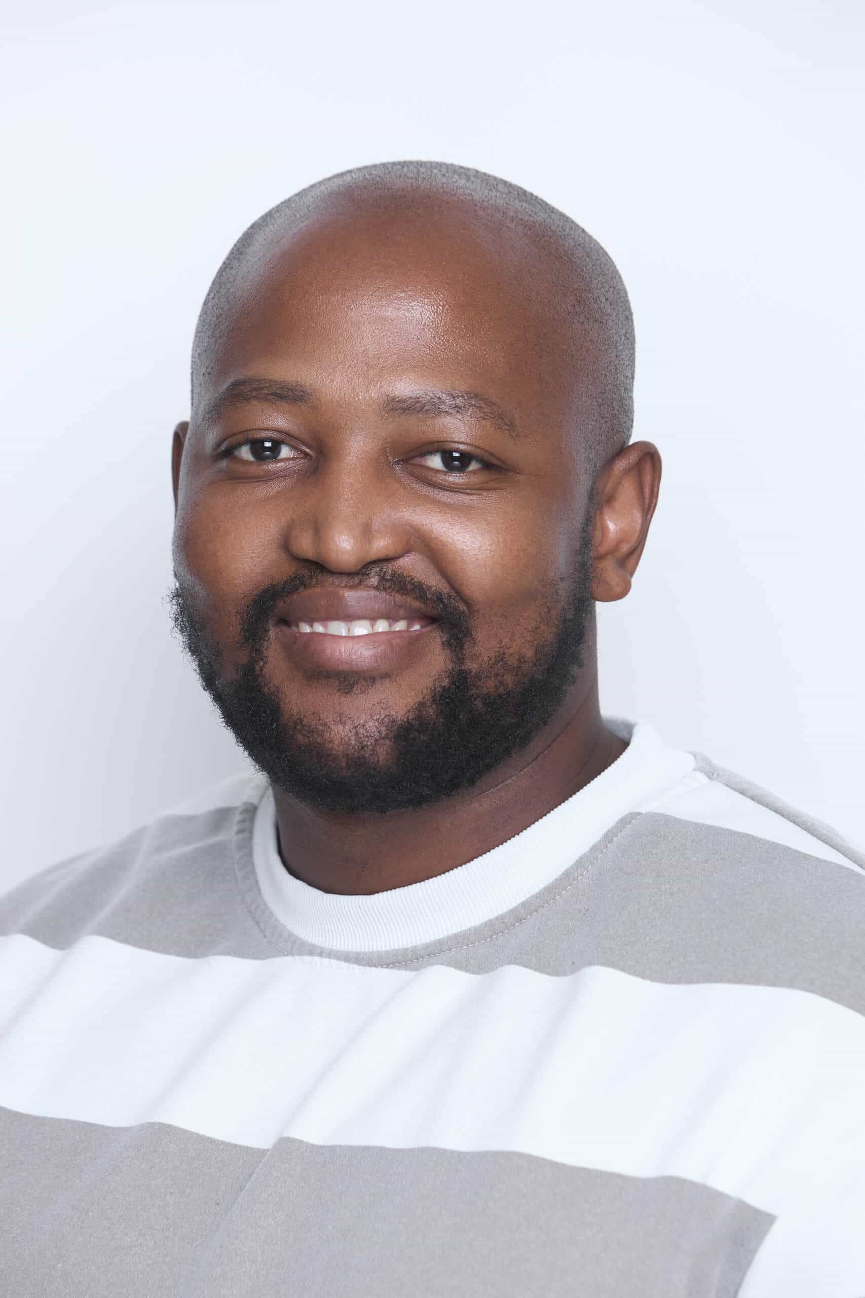 A man with a shaved head and a trimmed beard smiles at the camera. He is wearing a light gray and white striped sweater. The background is plain white, creating a clean and simple portrait setting.