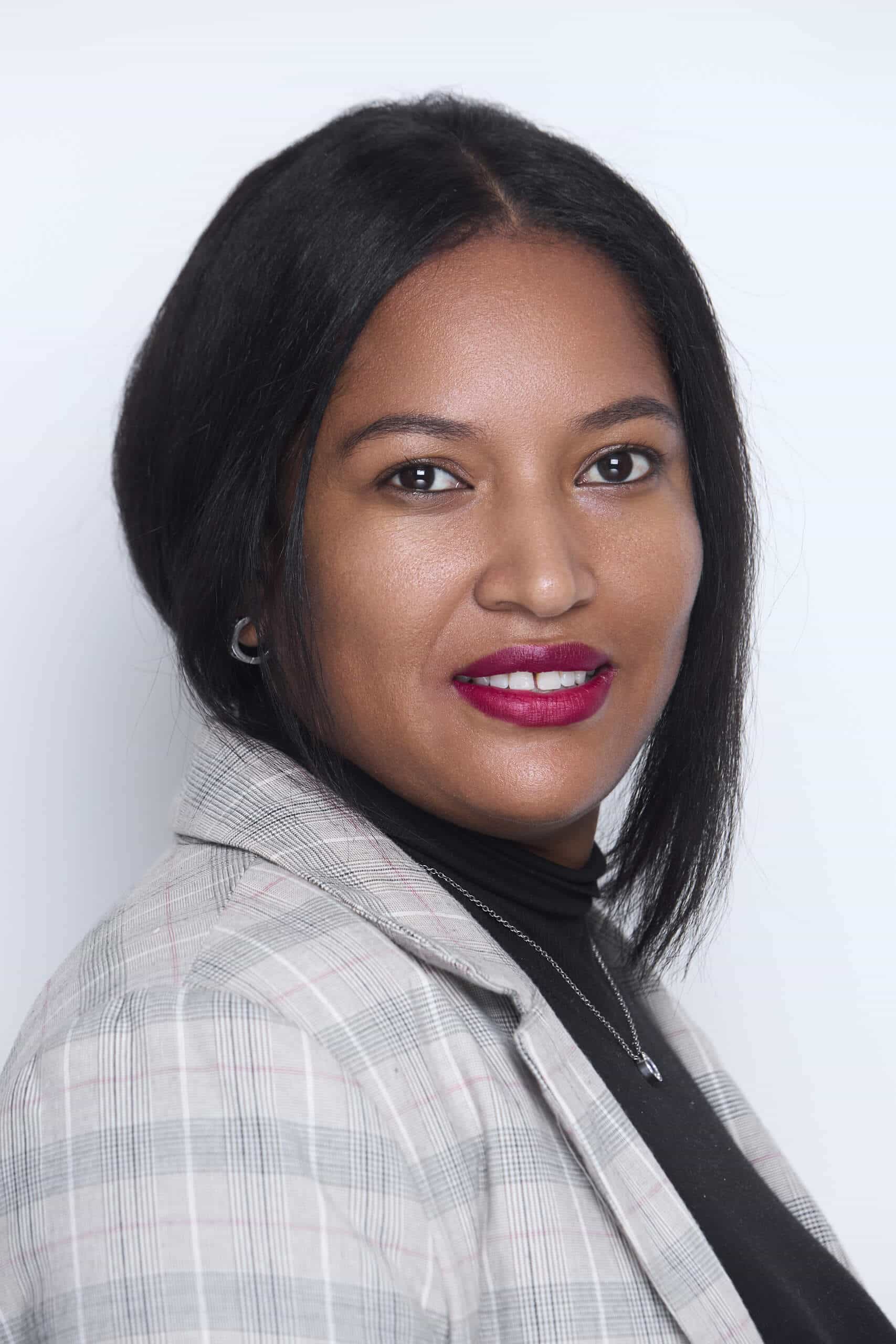 A woman with straight, shoulder-length black hair wears a light plaid blazer over a black turtleneck. She has medium brown skin, bold dark red lipstick, subtle makeup, and small hoop earrings. She faces the camera with a slight smile against a plain light background.