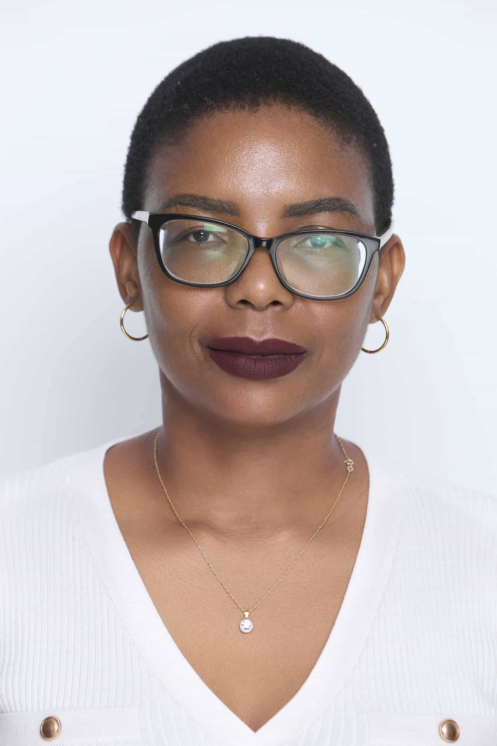 A person with short black hair wears glasses, gold hoop earrings, a gold necklace with a round pendant, and a white top with buttons. They have dark lipstick and appear in front of a plain, light background, facing the camera with a neutral expression.
