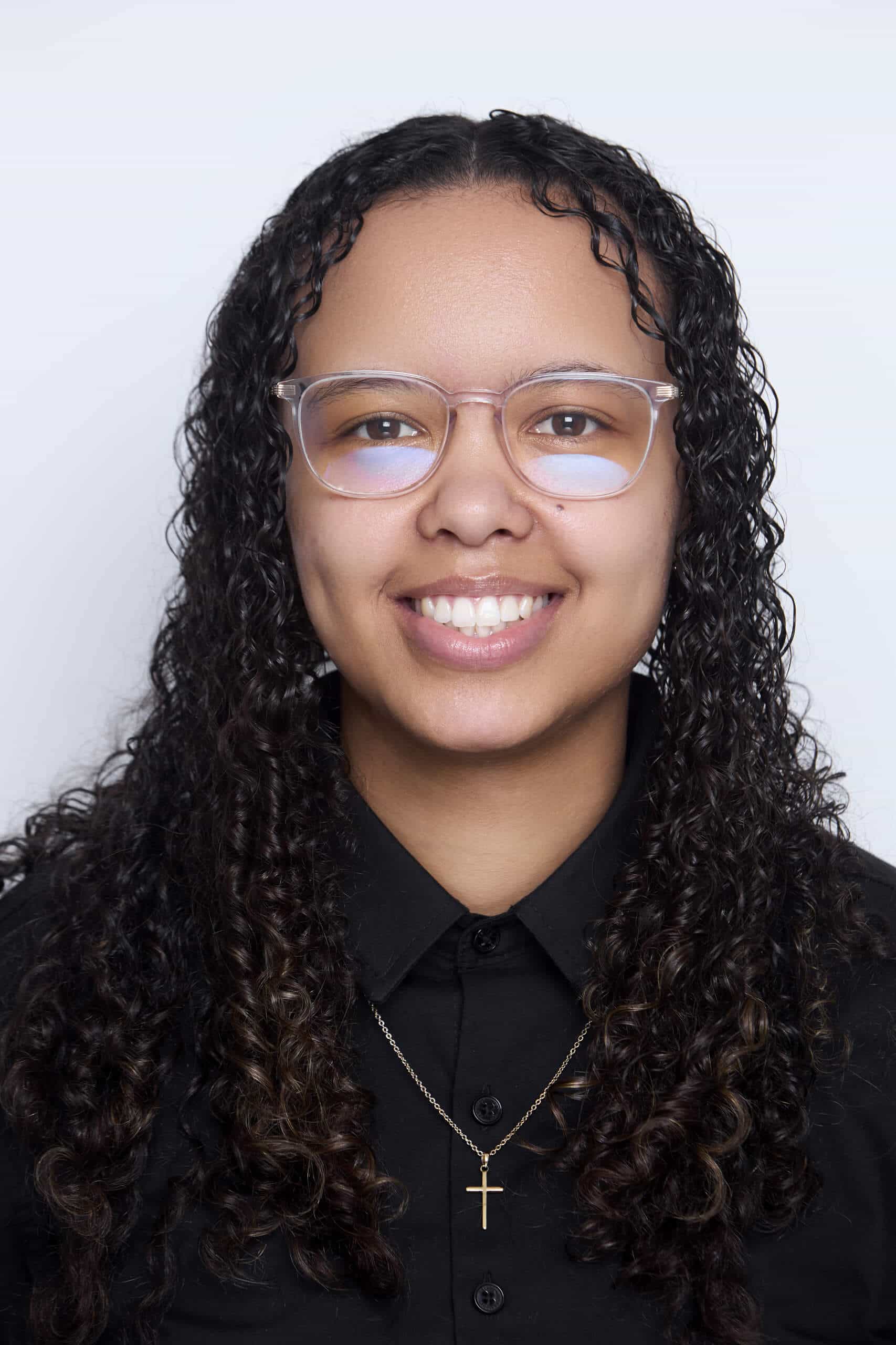 A person with medium brown skin and long, curly dark hair smiles at the camera. They wear clear glasses, a black collared shirt, and a gold necklace with a cross pendant against a plain white background.