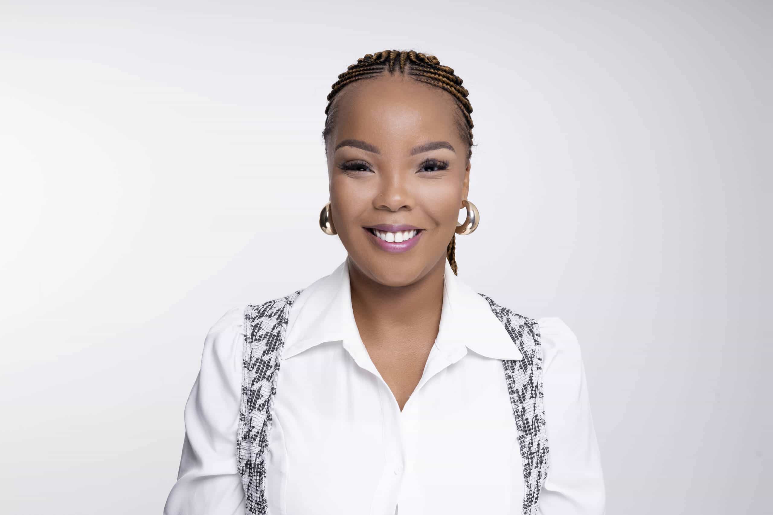 A woman with braided hair smiles at the camera. She wears gold hoop earrings, a white collared shirt, and a patterned vest. The background is plain white, giving the portrait a clean, professional look.
