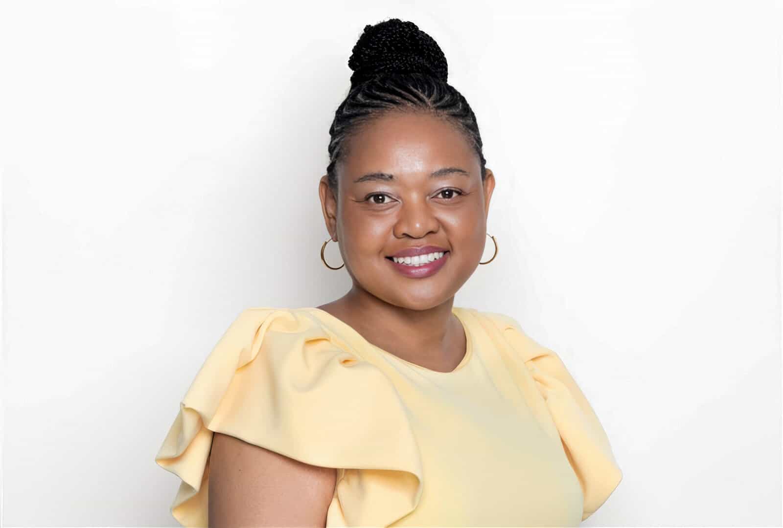 A woman with braided hair styled in a bun smiles at the camera. She wears gold hoop earrings and a light yellow top with ruffled sleeves. The background is plain white, creating a bright and clean portrait.