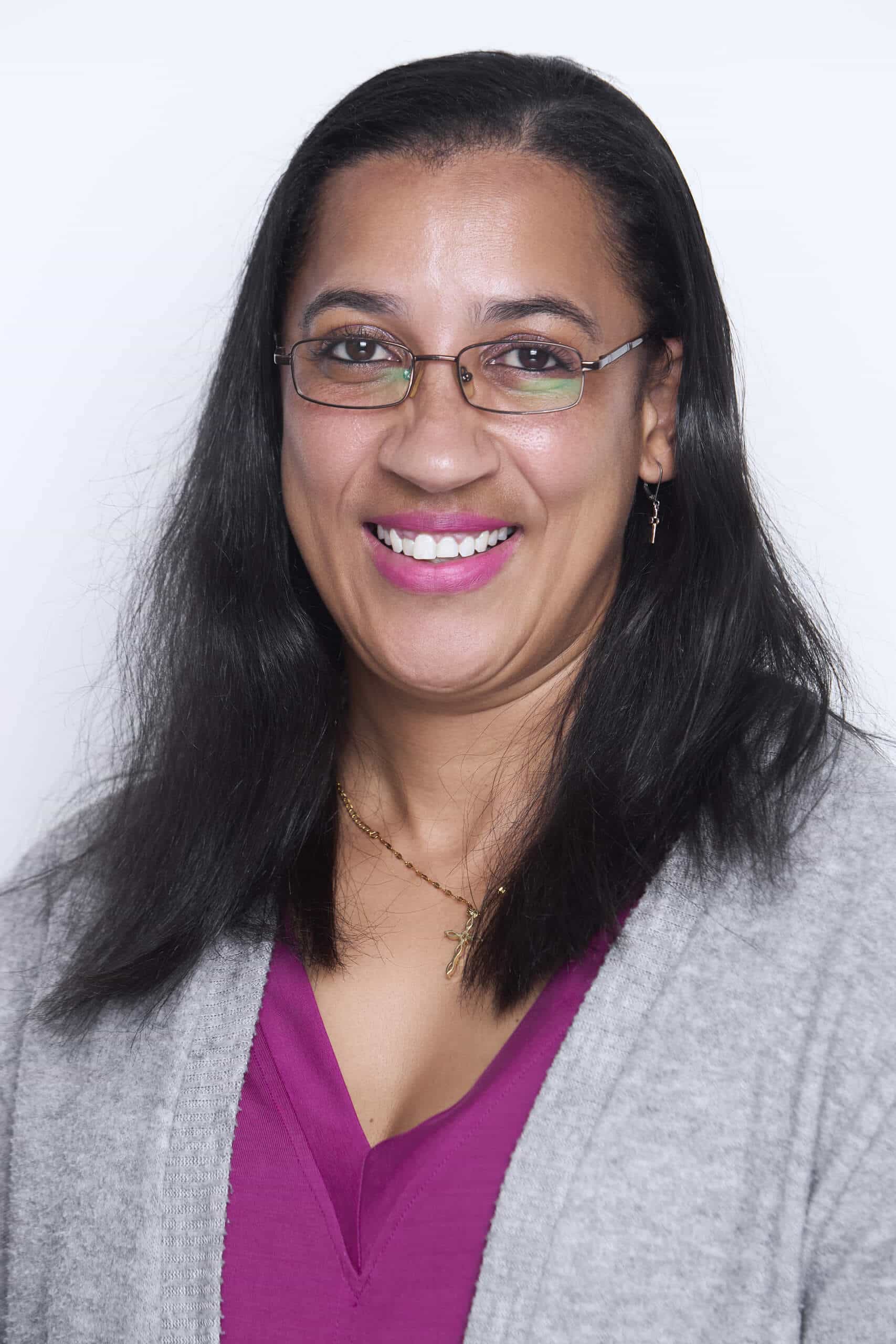 A woman with long dark hair, glasses, and medium skin tone smiles at the camera. She wears a magenta top, a light gray cardigan, small earrings, and two thin necklaces. The background is plain white.