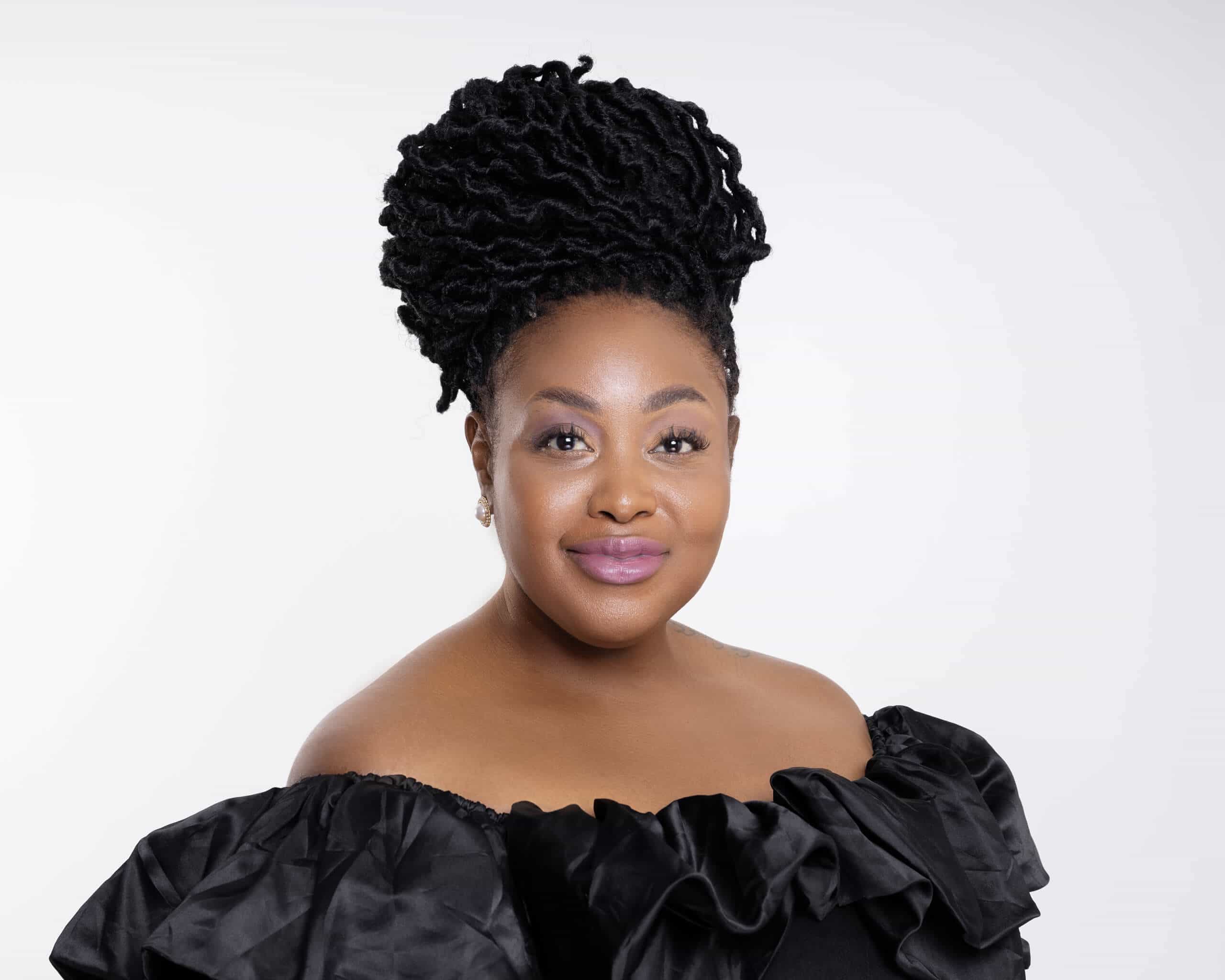 A woman with dark skin and long, styled black hair in an updo smiles softly. She wears a black, off-the-shoulder ruffled top and subtle makeup, with a light pink lip color. The background is plain white.