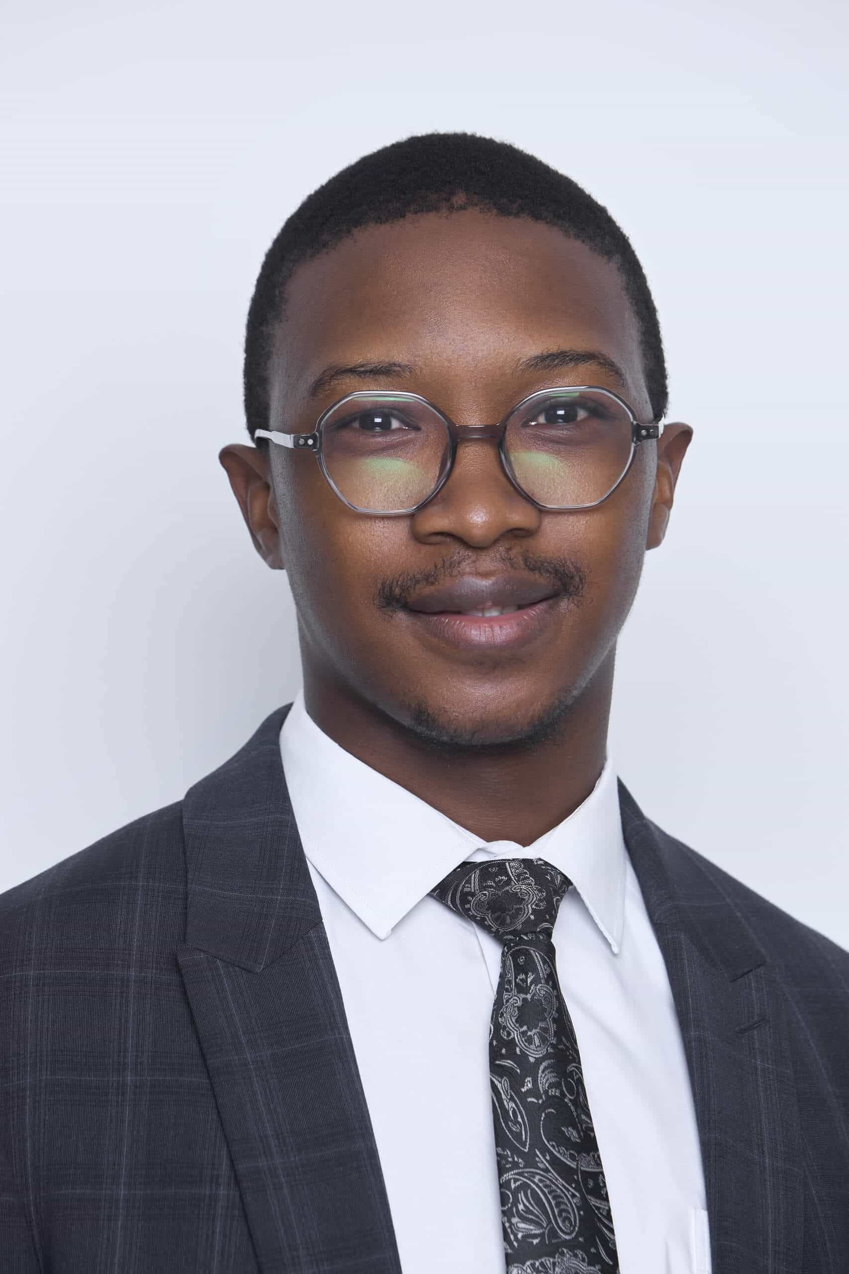 A man with short hair and glasses, wearing a dark plaid suit jacket, white dress shirt, and a black paisley tie, stands against a plain light gray background, smiling slightly at the camera.