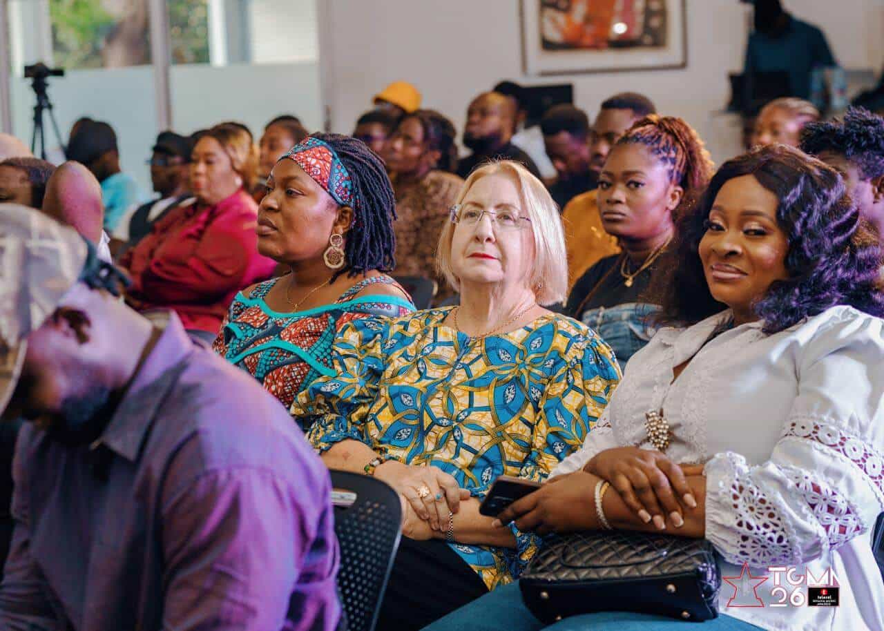 A diverse group of people, including women and men of different ages and backgrounds, sit closely together in rows, attentively watching an event. Most are focused forward; some smile, while others appear thoughtful or neutral. Indoor setting with bright lighting.