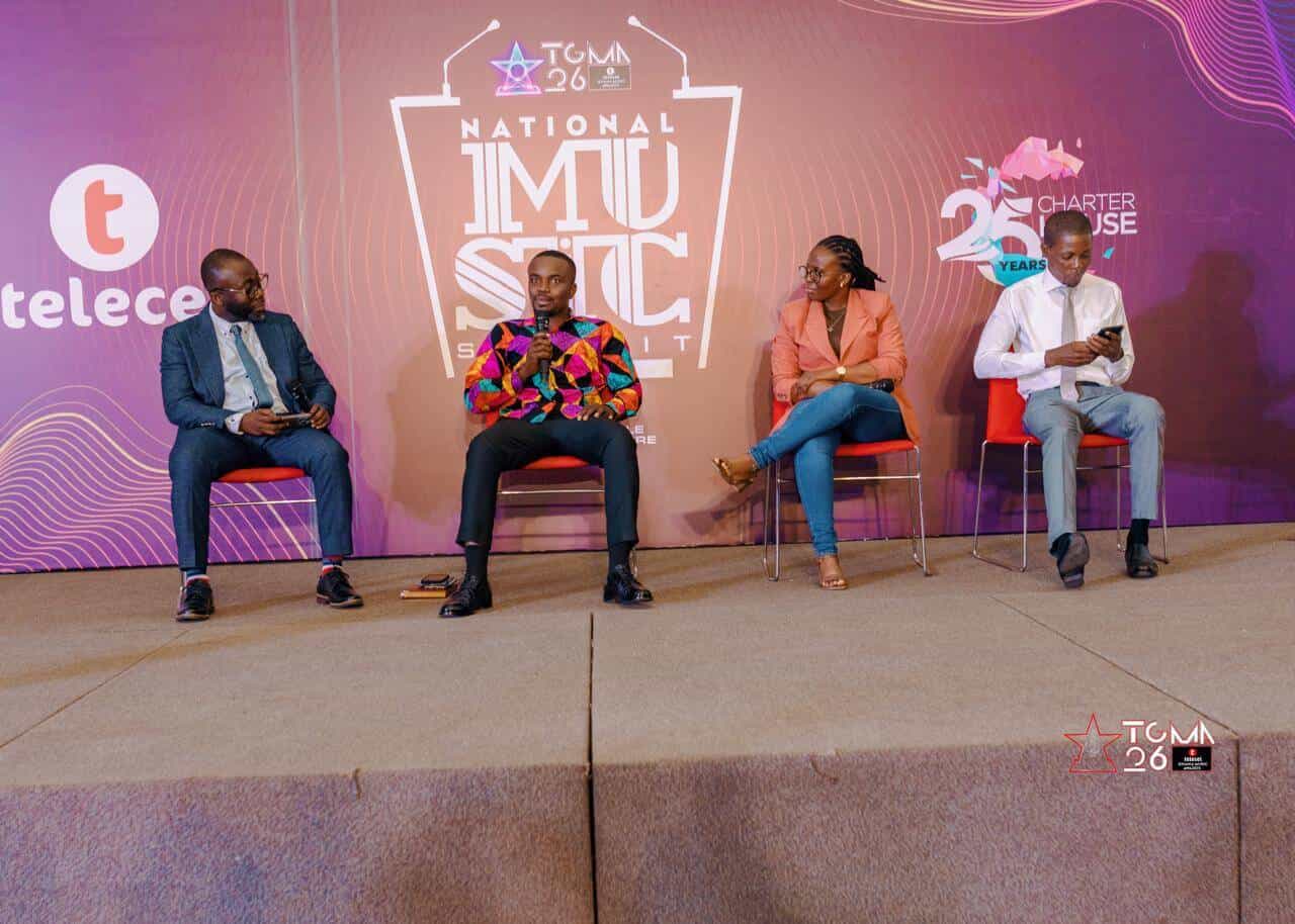 Four people sit spaced apart on red chairs on a stage at the National Music Summit. Behind them is a purple backdrop with event logos and branding. Two men wear suits, one man wears a colorful shirt, and a woman wears a pink jacket and jeans.