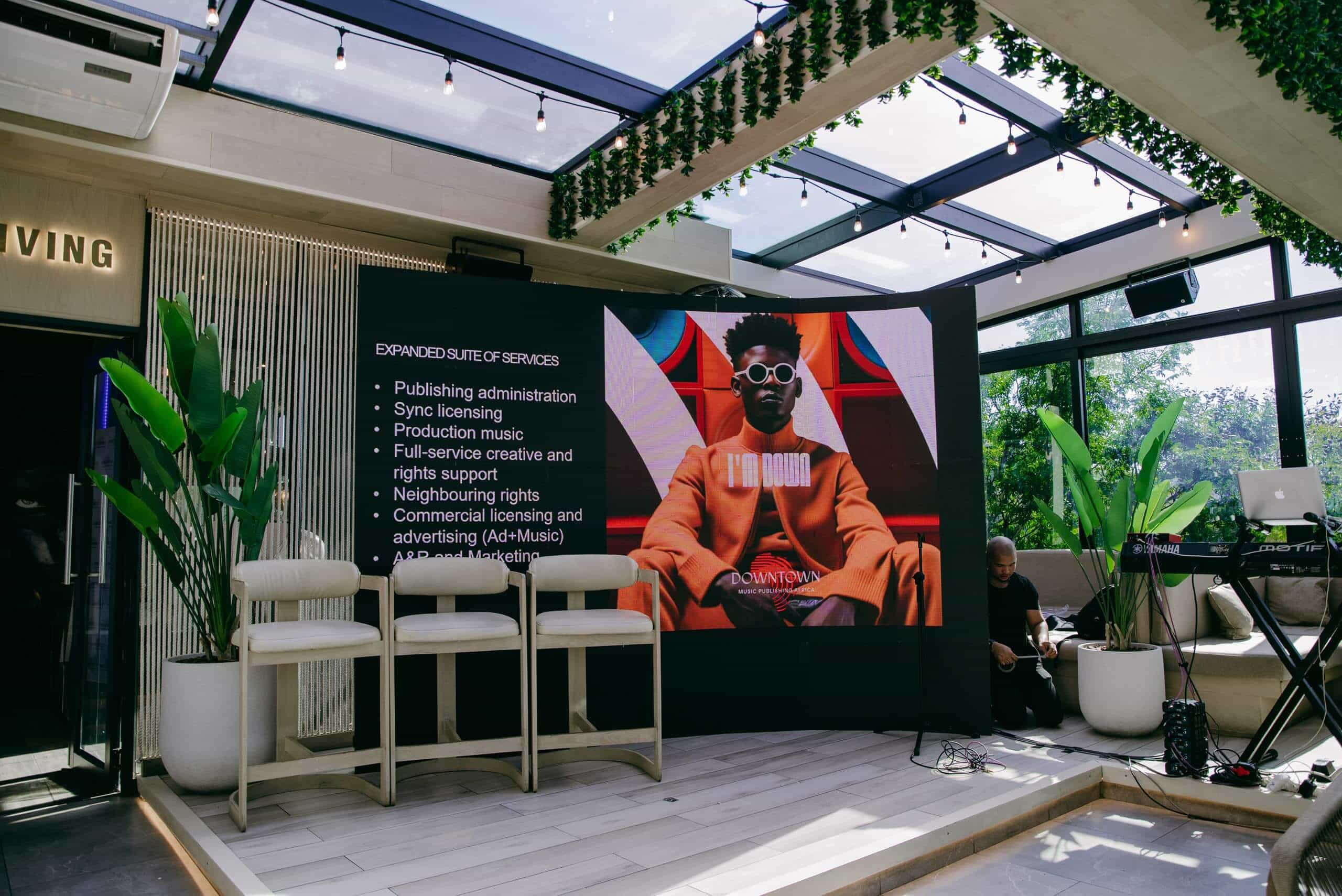 A modern indoor event space with a large digital screen displaying a man in an orange suit and sunglasses. The screen lists music industry services. Three white chairs sit on stage; a keyboard is on the right, with leafy plants and natural light filling the room.