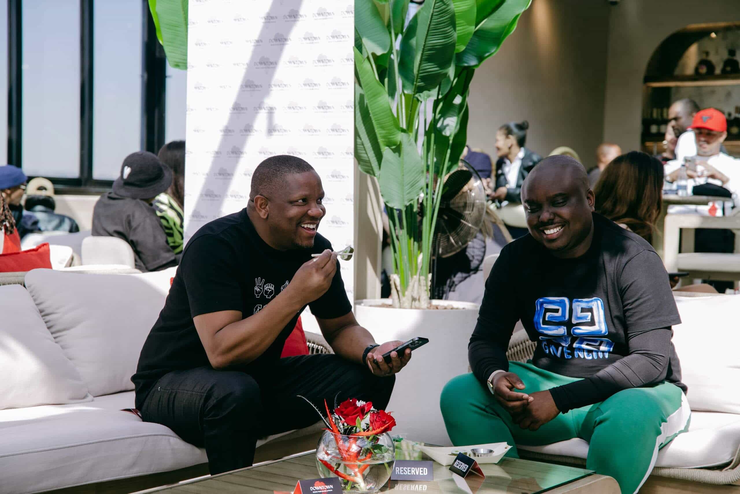 Two men sit on a white outdoor sofa, smiling and laughing. One holds a spoon and a phone, the other wears a black shirt with blue-green graphics. A &ldquo;RESERVED&rdquo; sign and a bowl with red flowers are on the table. Green plants and people appear in the background.