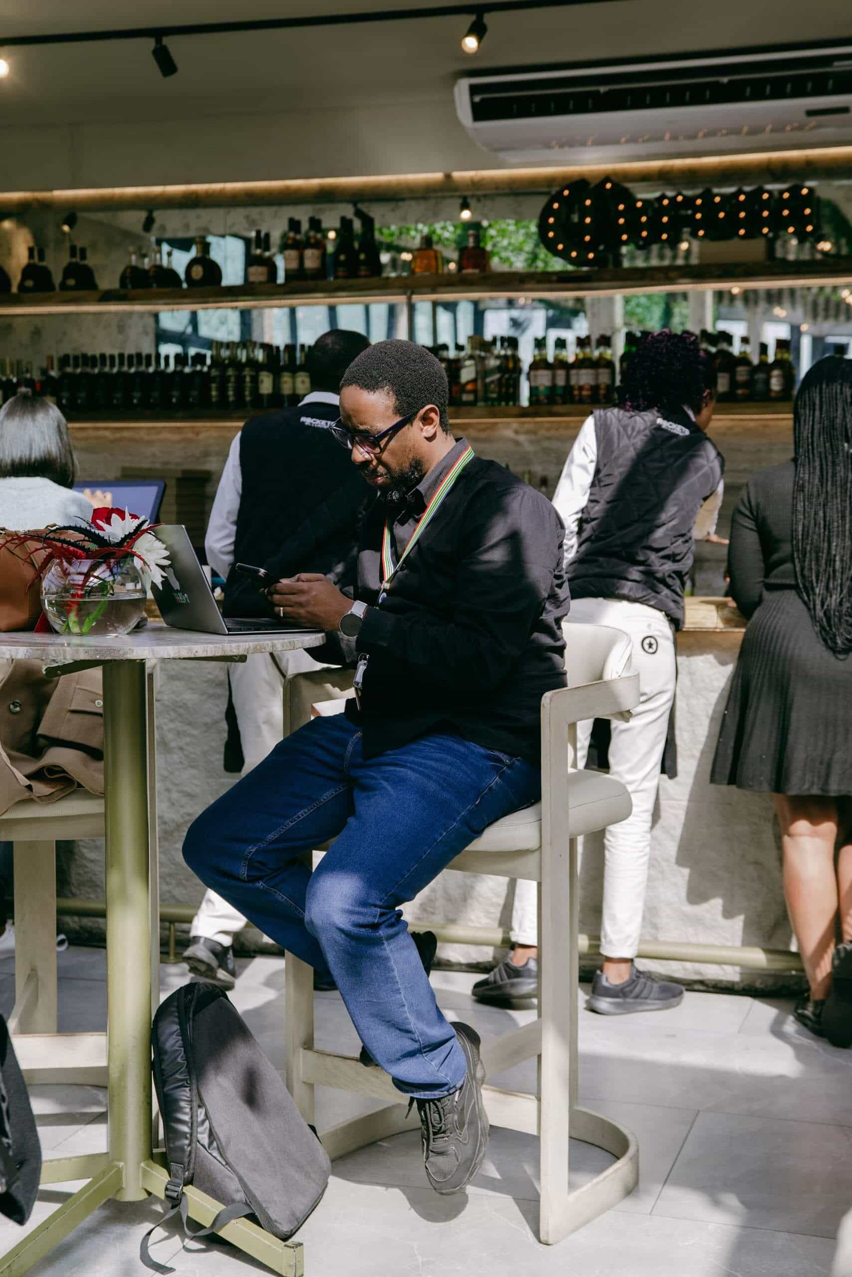 A man in glasses sits on a high chair at a bar, using a laptop and phone. He wears a black sweater, blue jeans, and black shoes. A black backpack is on the floor near his feet. Other patrons and bartenders are visible in the background.