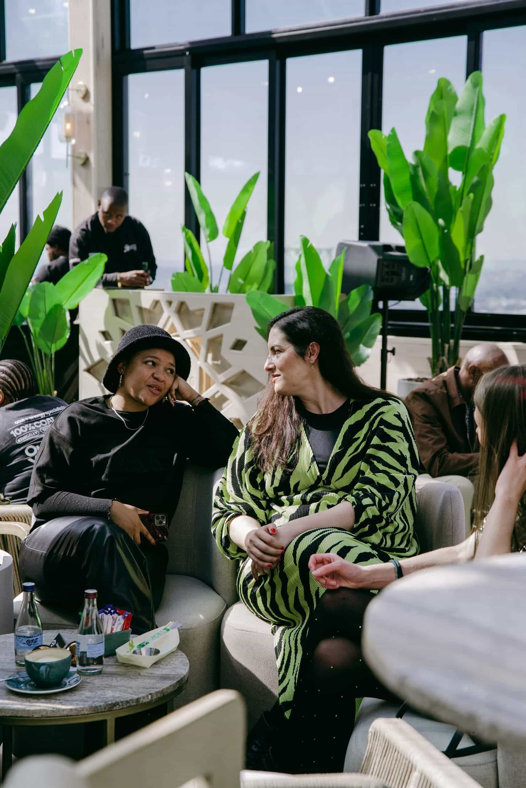 Two women sit and talk on modern lounge chairs in a bright, plant-filled indoor space. One wears a black outfit and hat, the other wears a green and black animal-print dress. Other people and tables with drinks and snacks are in the background.