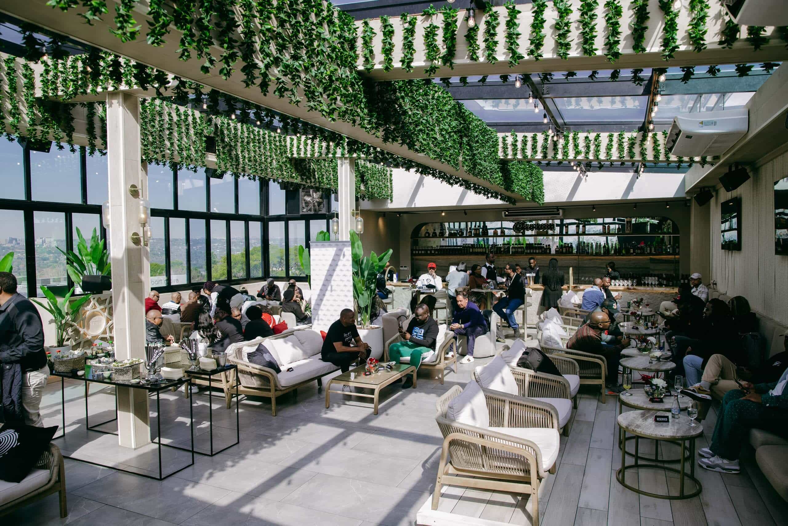A lively rooftop lounge with many people sitting on sofas and chairs, chatting and enjoying drinks. The space has greenery hanging from the ceiling, large windows, potted plants, and a bar at the back. Sunlight streams in through the glass roof.