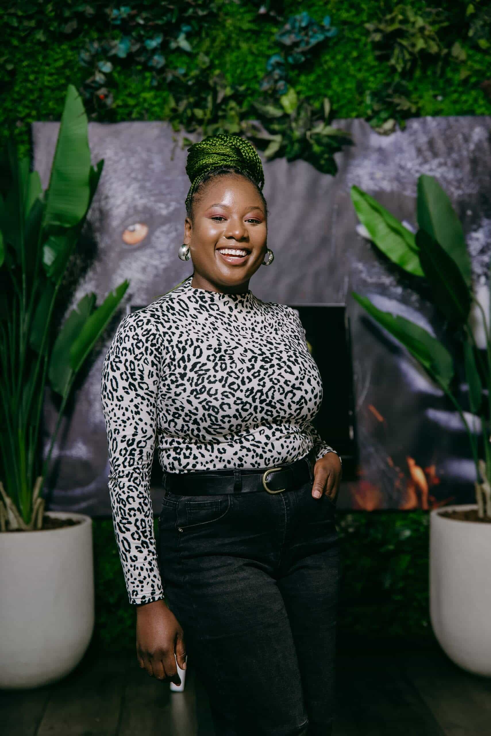 A woman with green braided hair styled in a bun stands smiling indoors. She wears a black-and-white leopard print top, black jeans, and hoop earrings. Behind her are potted plants and a dark leafy backdrop. She holds a phone in her left hand.