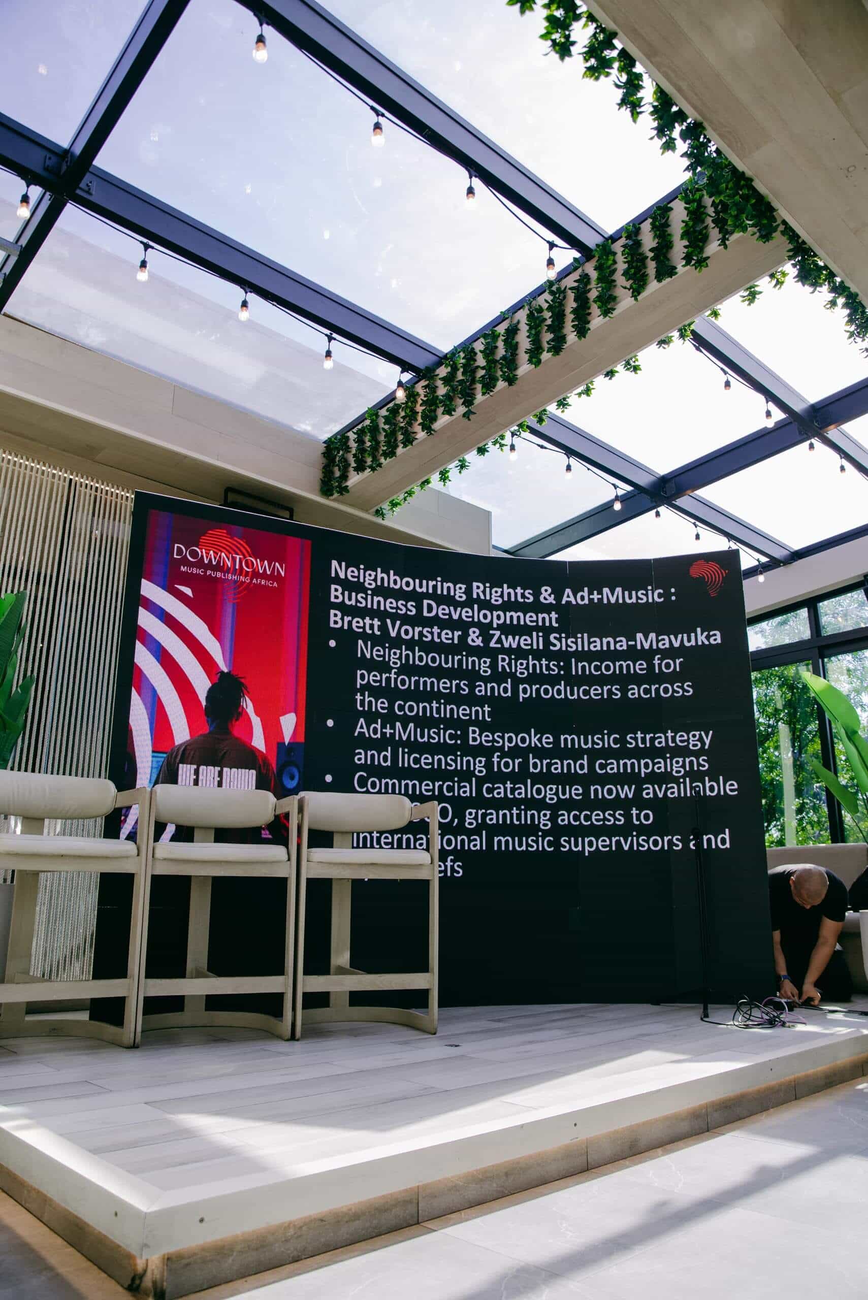 A modern indoor event space with a large digital screen displaying information about music rights. There are two high stools in front of the screen, plants nearby, and a person crouching on the right arranging cables. The ceiling has glass panels and greenery.