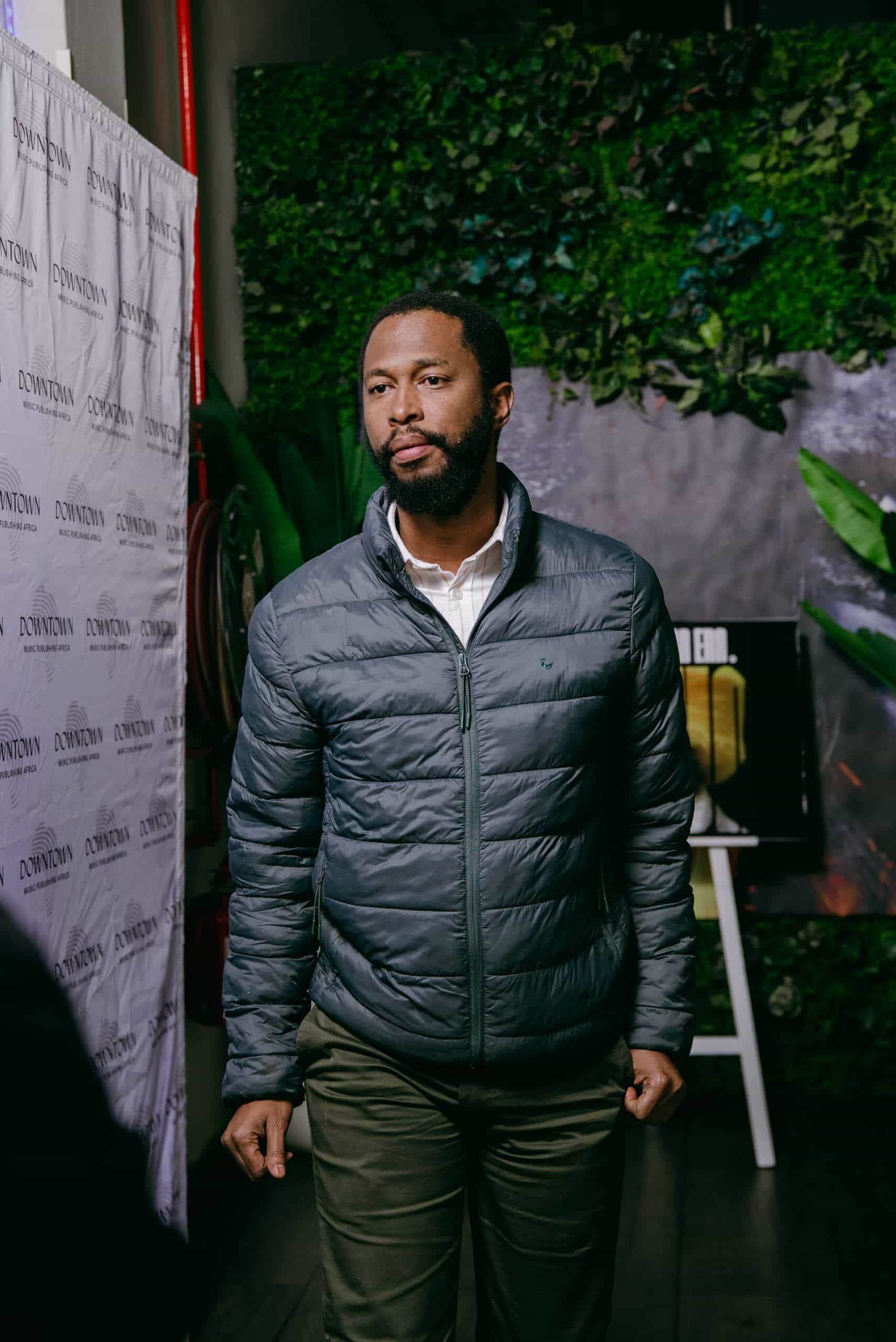 A man with a beard wearing a gray puffer jacket and olive pants stands indoors. Behind him is a green wall with plants, a poster, and a white step-and-repeat banner with &ldquo;DOWNTOWN&rdquo; written repeatedly. He looks thoughtful, with hands by his sides.