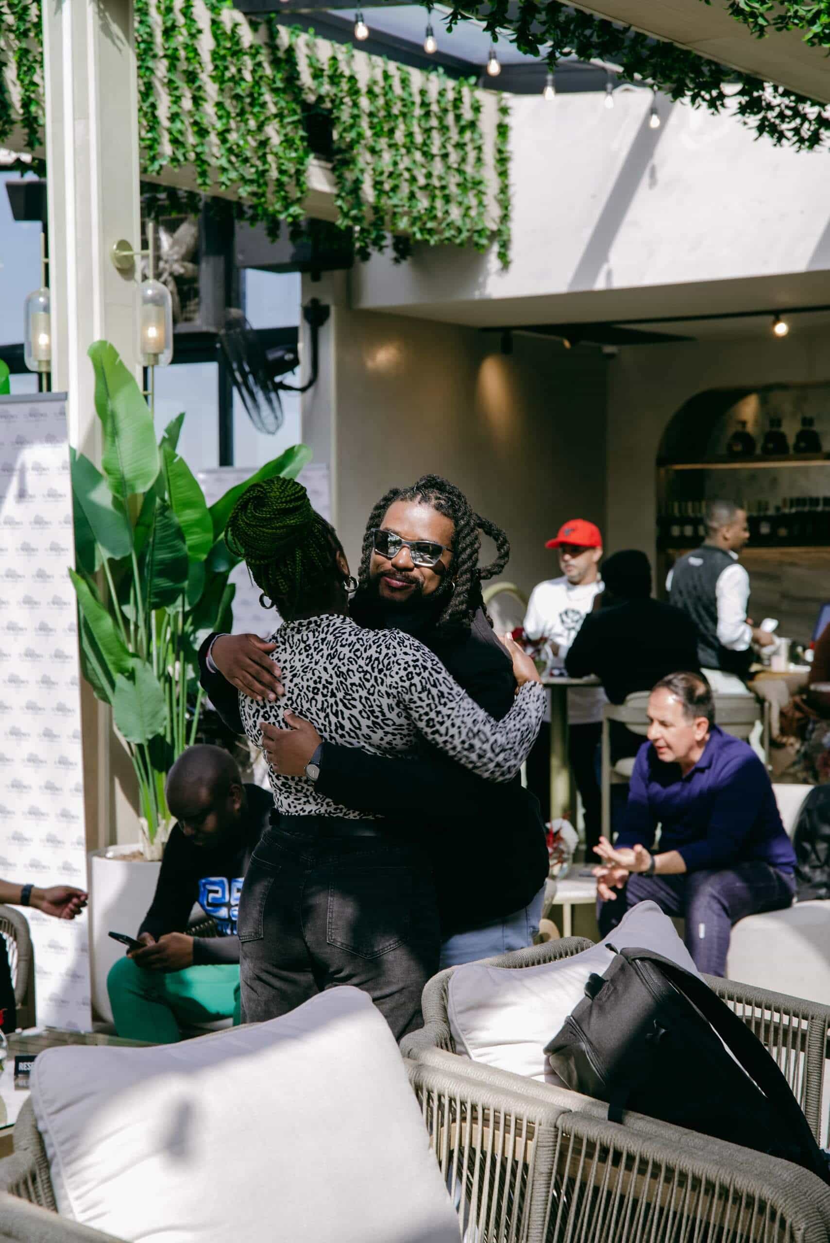 Two people embrace and smile in a stylish outdoor lounge with plants and cozy seating. Other people sit and talk in the background, some using phones. Sunlight brightens the scene, and a leafy decor hangs above. The mood is friendly and relaxed.