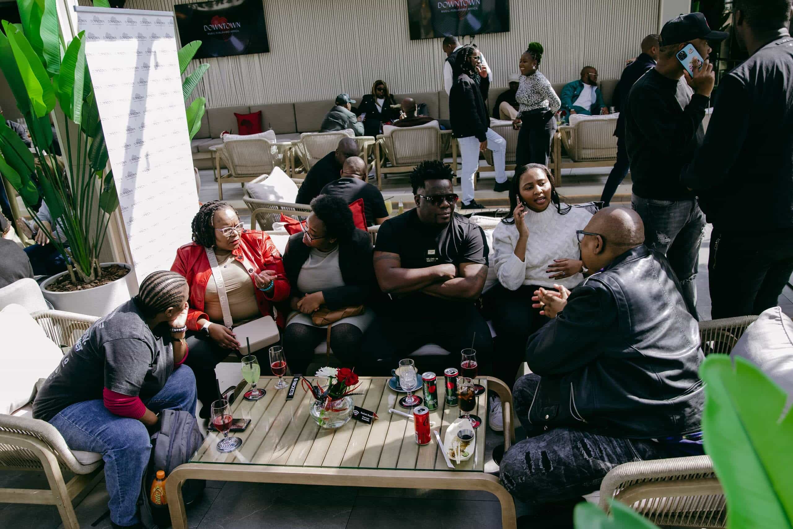 A group of people sit around a light wooden table outdoors, chatting and laughing. The table holds drinks, snacks, and flowers. Other people are socializing in the background. Green plants and couches are visible, creating a lively, relaxed atmosphere.