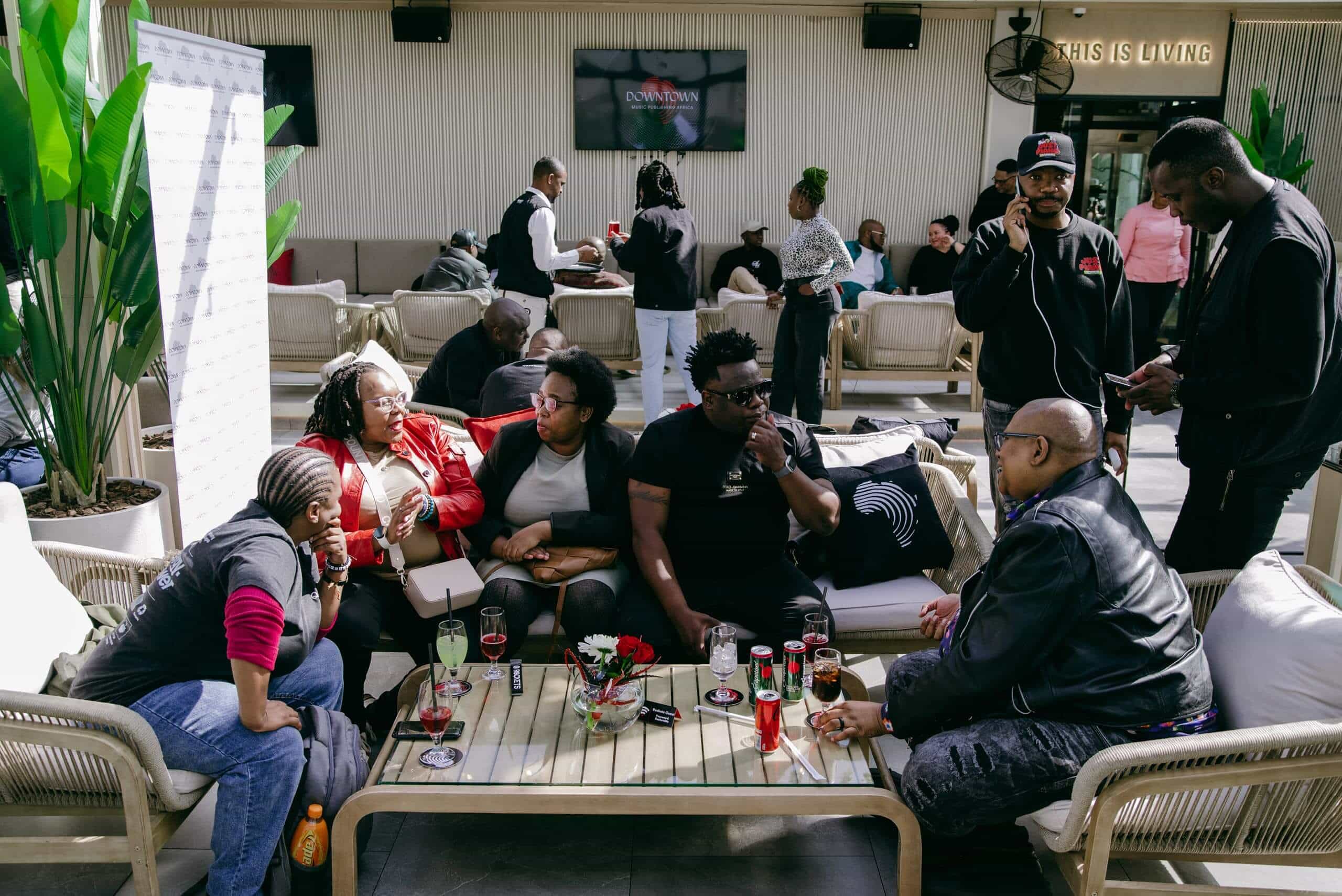 A group of people sit and stand around a low outdoor table, chatting and enjoying drinks. The area has cushioned seats, plants, and people in the background. Glasses, cans, and snacks are on the table, creating a lively, social atmosphere.