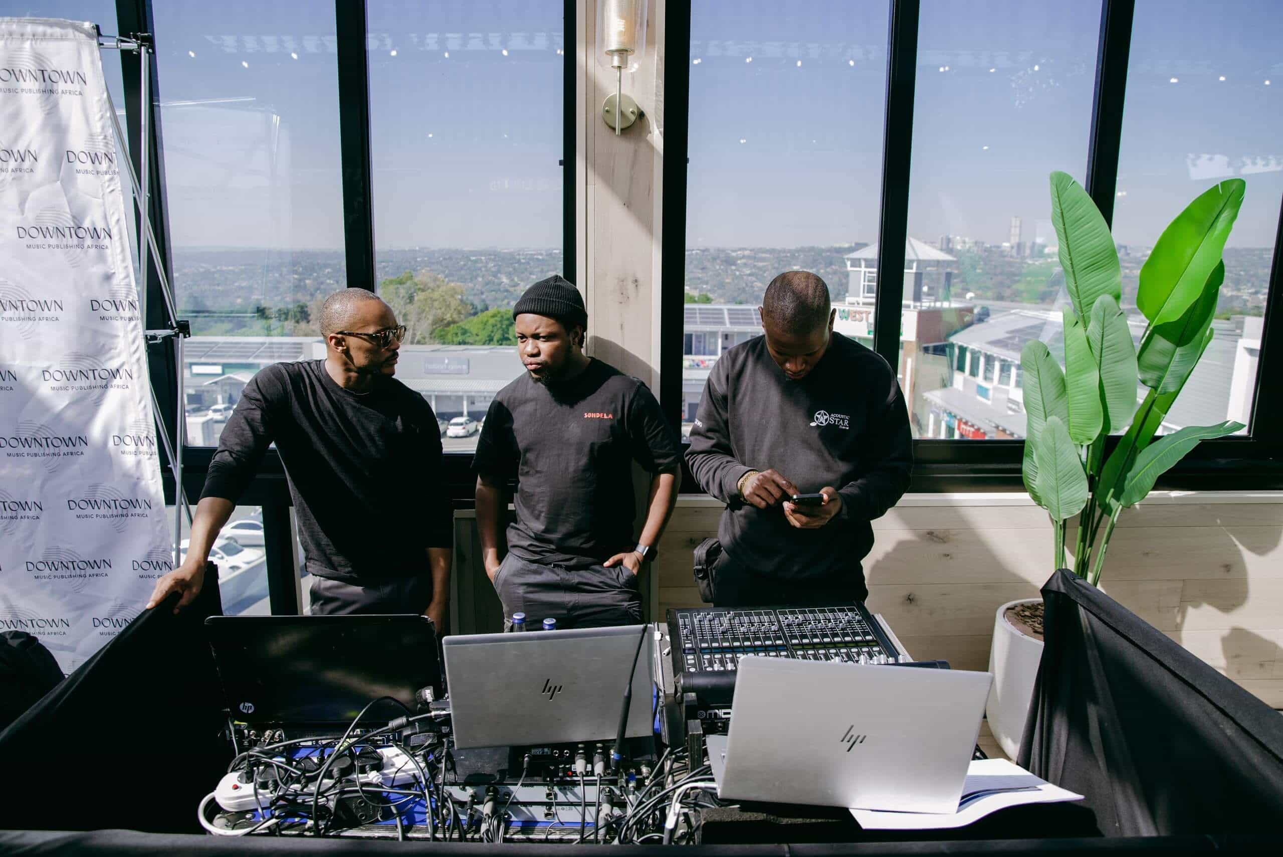Three men stand behind a DJ booth with laptops, sound equipment, and audio mixer. Two are talking while the third looks at his phone. Large windows reveal a cityscape and sky. A tall potted plant and event backdrop are visible in the bright modern room.