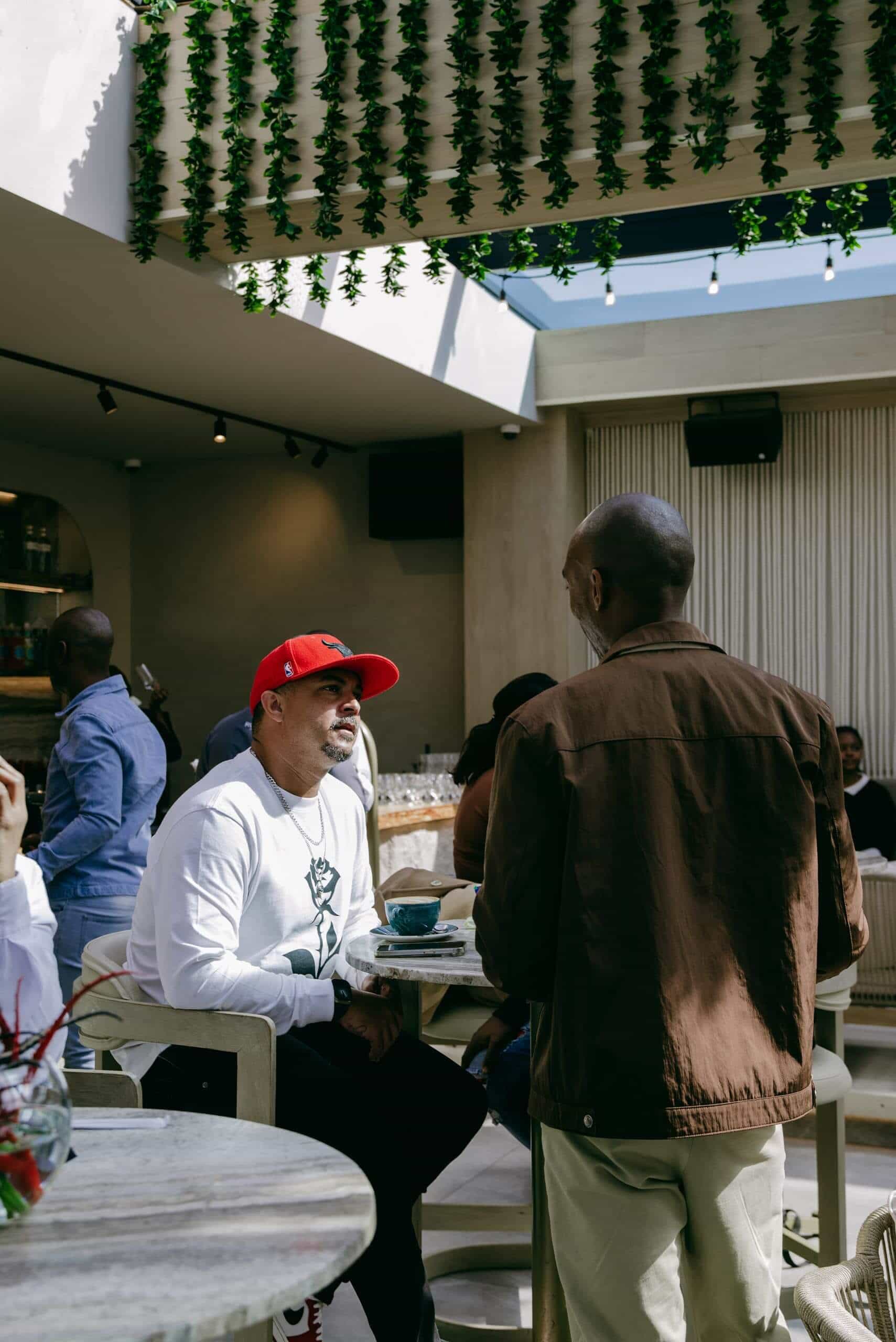 Two men converse at a caf&eacute; table. One wears a red cap and white sweatshirt with a black rose print, sitting with a drink. The other stands, wearing a brown jacket. Greenery hangs overhead; others are visible in the modern, sunlit space.