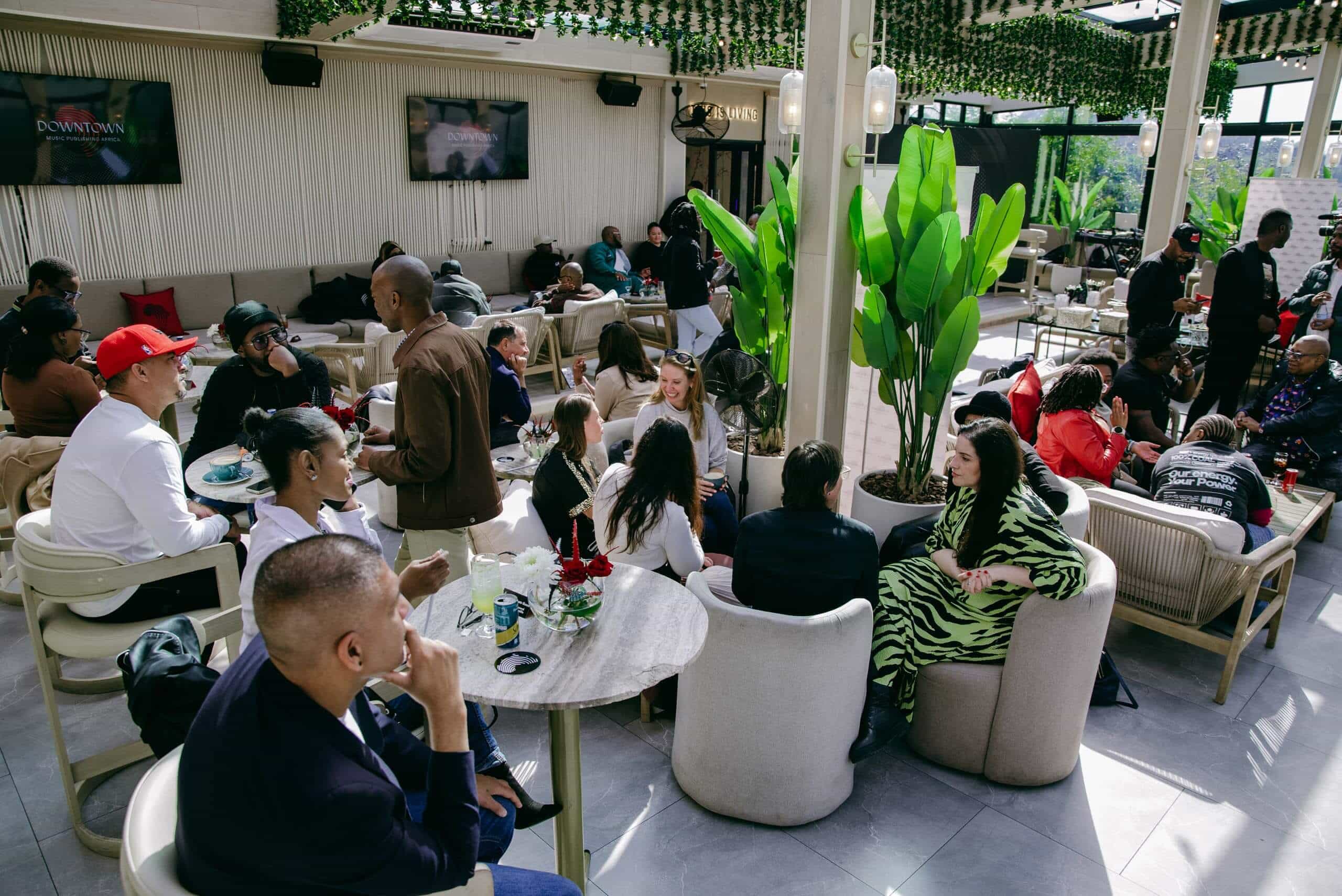 A lively group of people sits and chats at tables in a bright, modern lounge with green plants and large windows. Some are eating or drinking. The mood appears social and relaxed, with a mix of casual and trendy attire. Two TVs hang on the wall in the background.