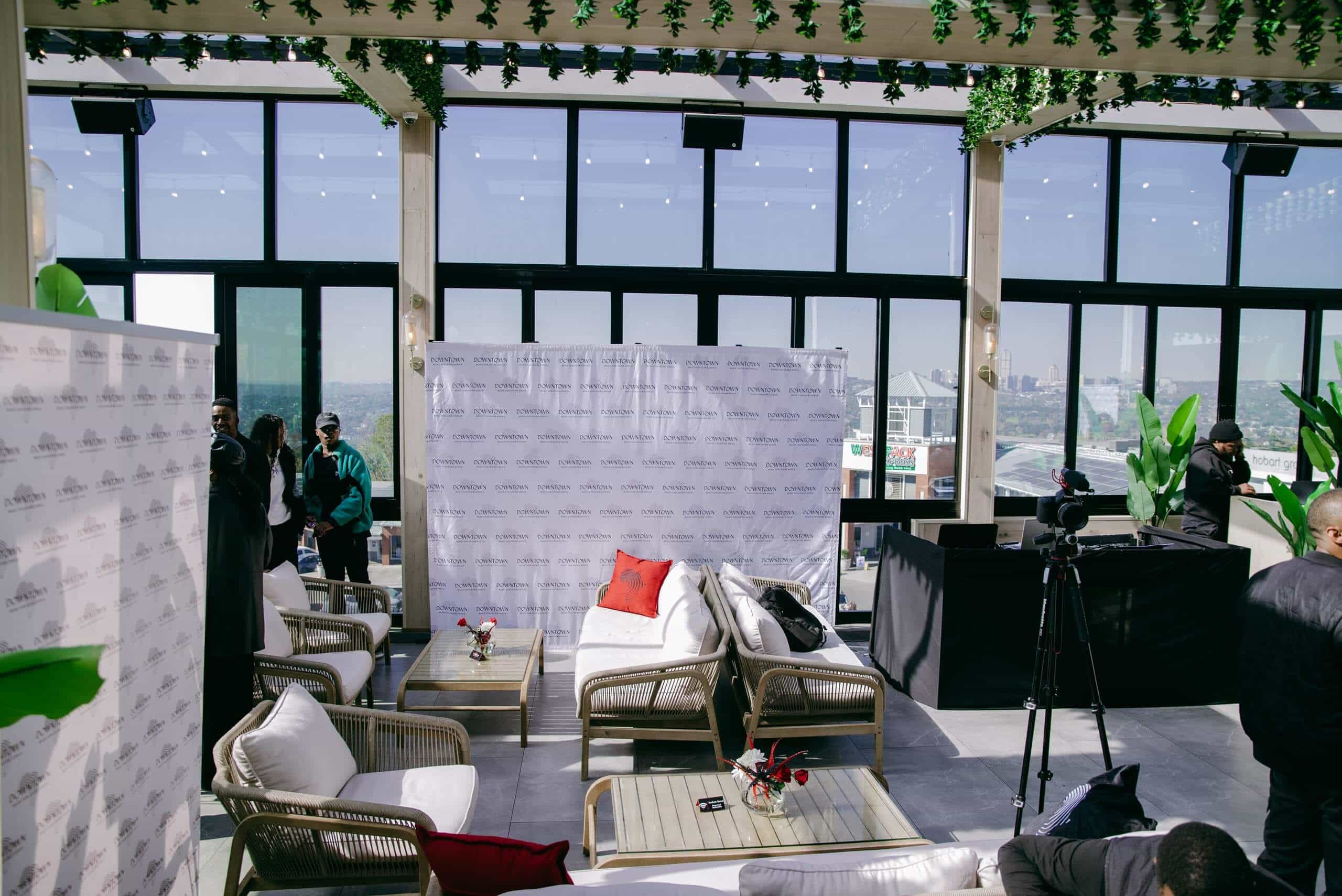 A modern rooftop lounge with wicker chairs, a white sofa, red and white cushions, and small flower vases. Greenery hangs above. A white step-and-repeat banner stands near large windows overlooking a city. Several people and cameras are visible on the left and right.