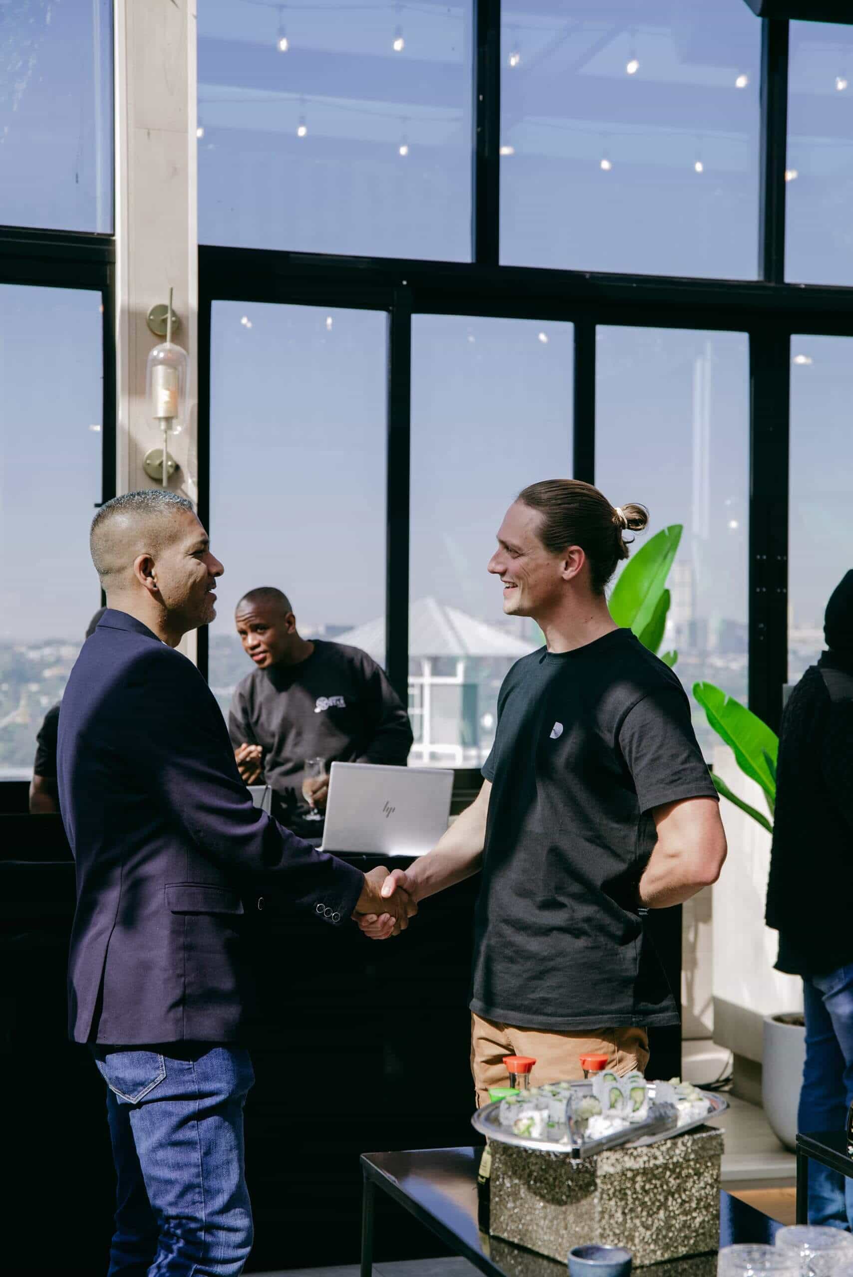 Two men shake hands and smile in a modern indoor space with large windows and potted plants. One wears a dark blazer, the other a black T-shirt and tan pants. In the background, a person works at a laptop on a counter. Bright daylight fills the room.