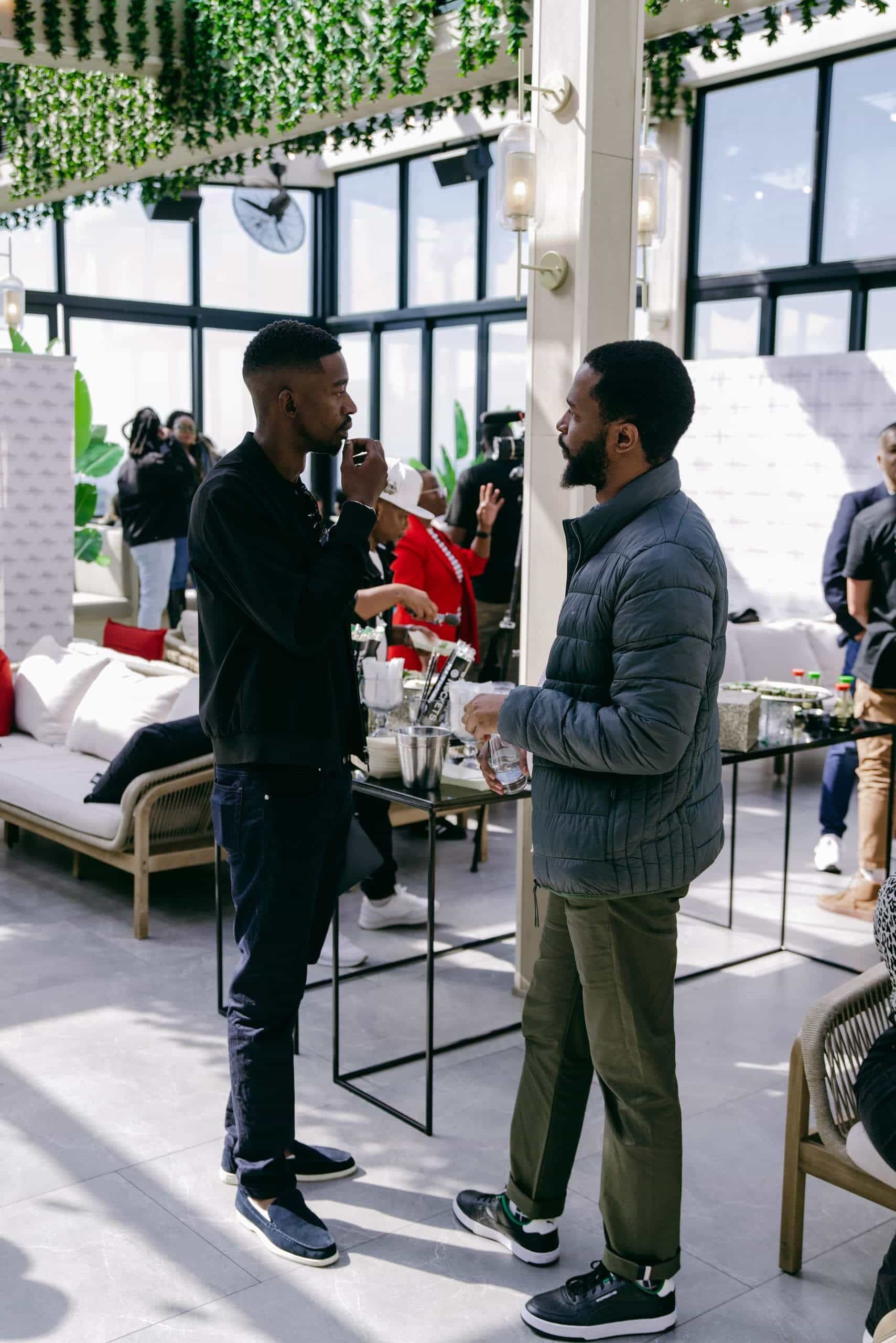 Two men stand and talk in a bright, modern indoor space with large windows and leafy decorations. One man eats while the other listens, and people mingle in the background near a table with drinks and snacks. The atmosphere appears social and relaxed.