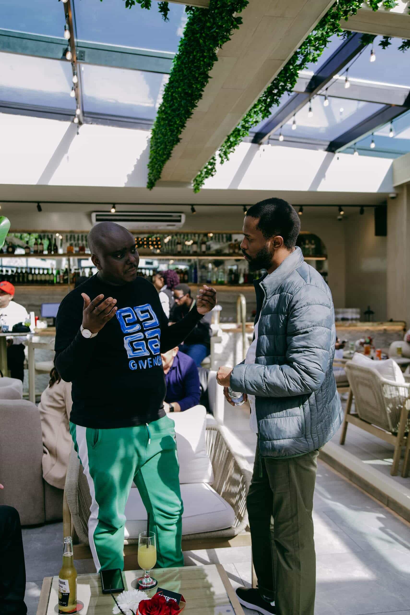 Two men stand and talk in a bright, modern indoor lounge with a glass ceiling and plants overhead. One man gestures expressively, wearing a black sweatshirt and green pants, while the other listens, wearing a gray jacket and khaki pants. People sit in the background.
