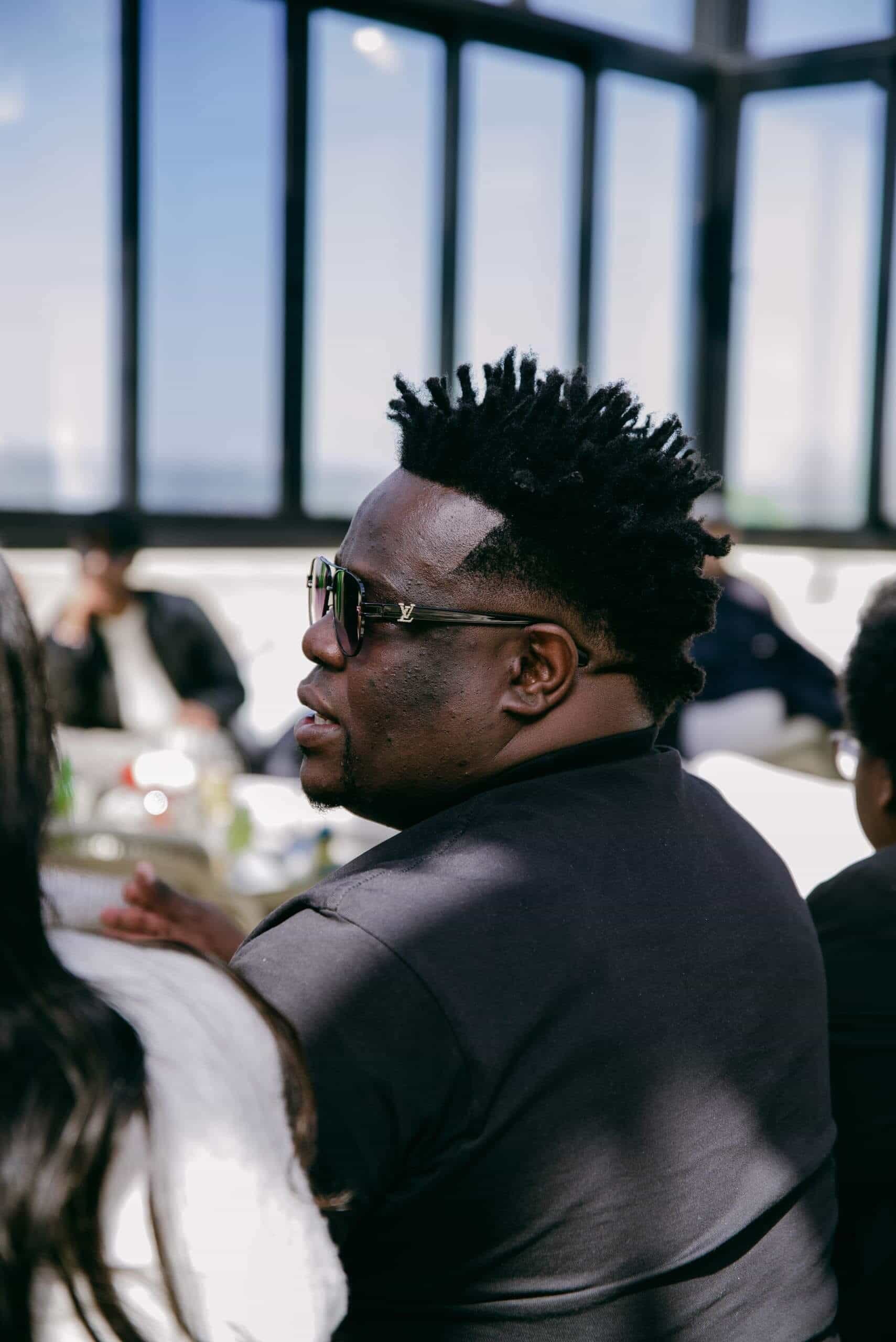 A man with short, spiked hair and sunglasses sits indoors, facing slightly to the left. He wears a dark shirt and appears to be listening to someone out of frame. The background shows large windows and blurred people sitting around a table.
