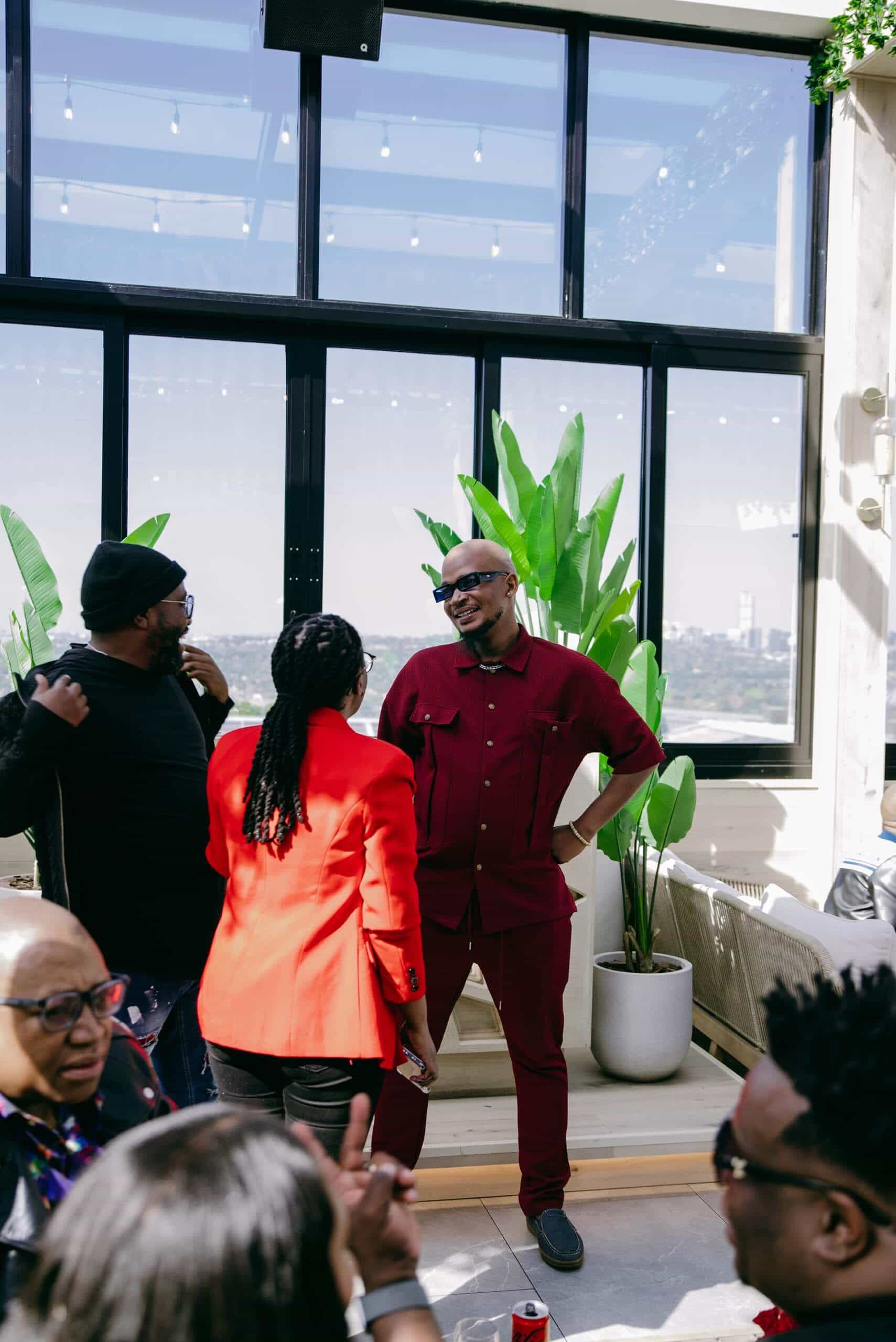 A man in a burgundy outfit and sunglasses stands, smiling with hands on hips, talking to a woman in a bright red blazer. Other people sit and stand nearby in a sunlit, modern indoor space with large windows and green plants.