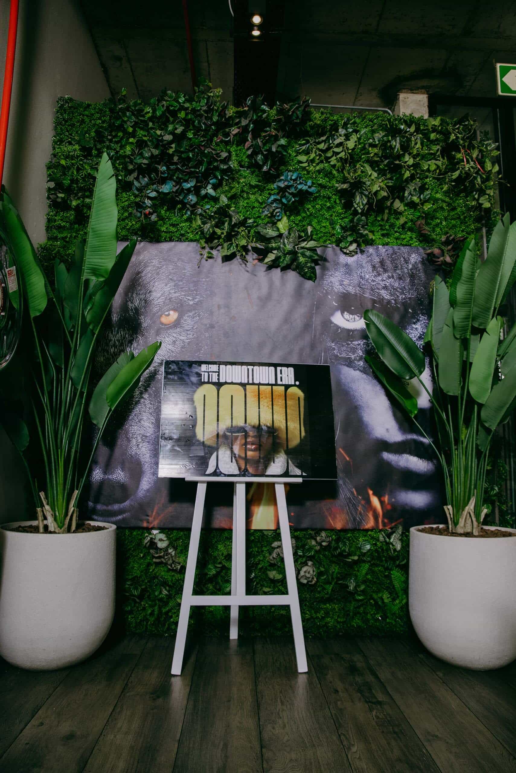 A poster on an easel stands before a backdrop of green foliage and large plant pots. The poster features a person's face overlaid with the word 