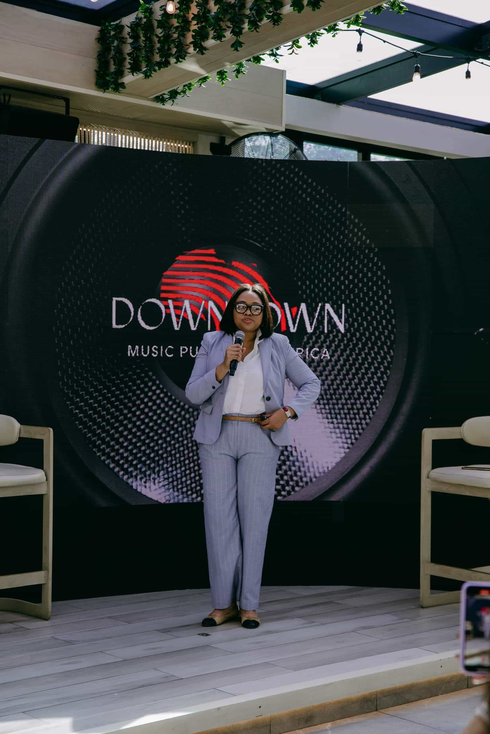 A woman in a light gray suit and glasses stands holding a microphone, speaking on stage in front of a large circular Downtown Music Publishing Africa logo, with two empty chairs on either side and greenery above.
