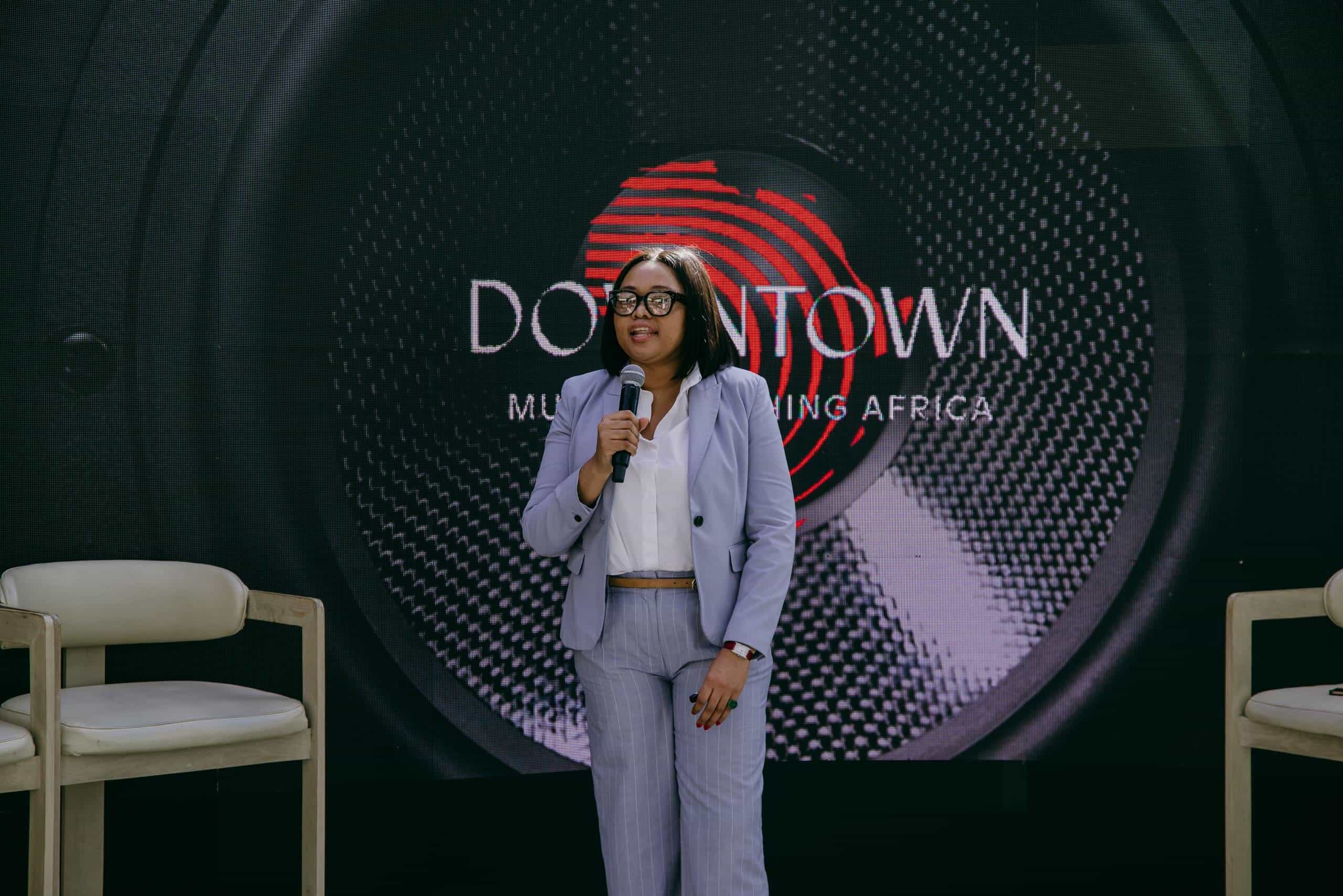 A woman in a light purple suit holds a microphone and speaks on stage. She stands between two empty chairs, in front of a large black backdrop displaying the 