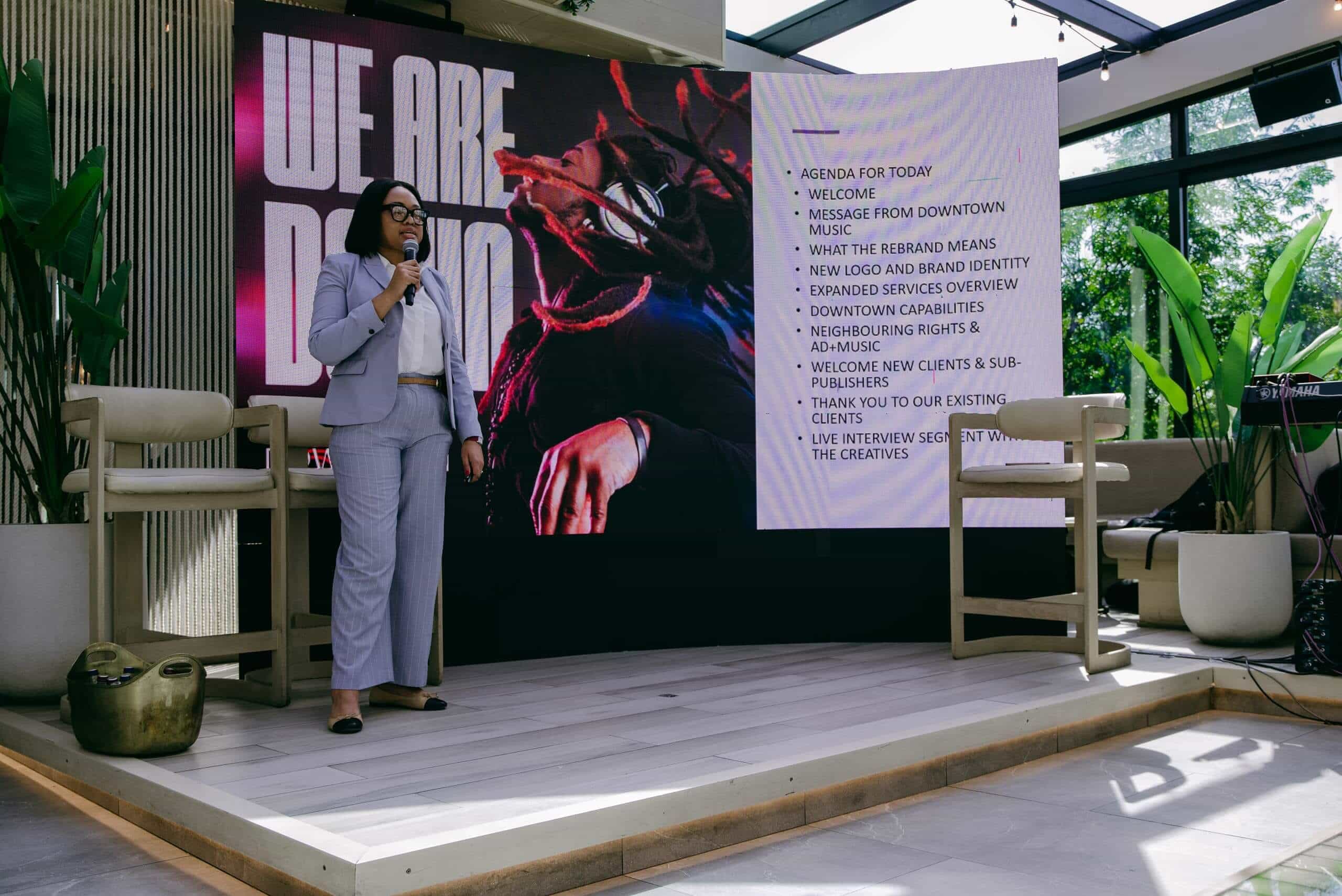 A woman in a gray suit speaks into a microphone on a modern indoor stage. Behind her, a large screen displays 