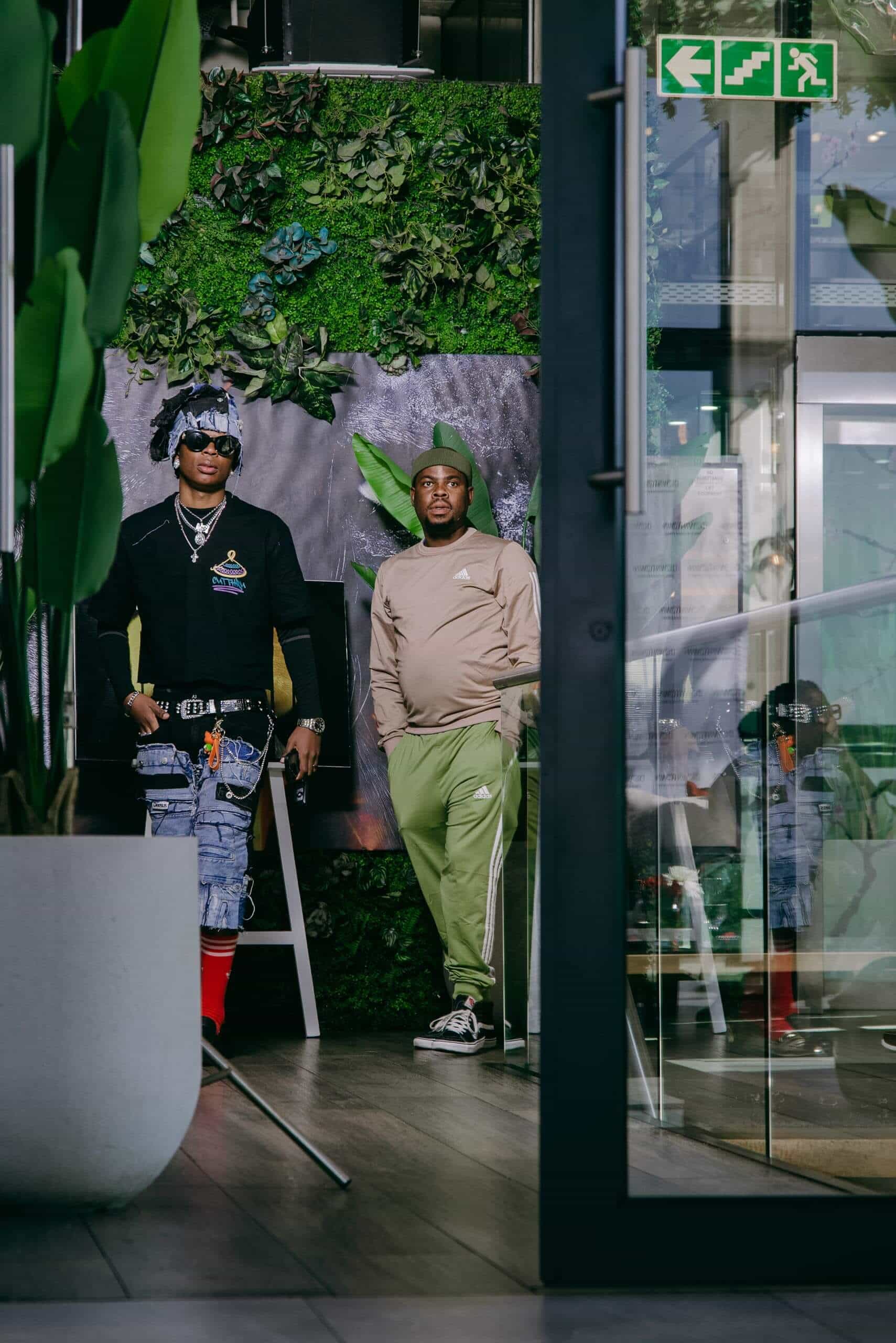 Two men pose indoors by a green, plant-covered wall. One wears a black cap, sunglasses, a black shirt, and blue jeans with red socks; the other wears a green tracksuit and beanie. Tall plants and a glass door are in the foreground, and an exit sign is above them.